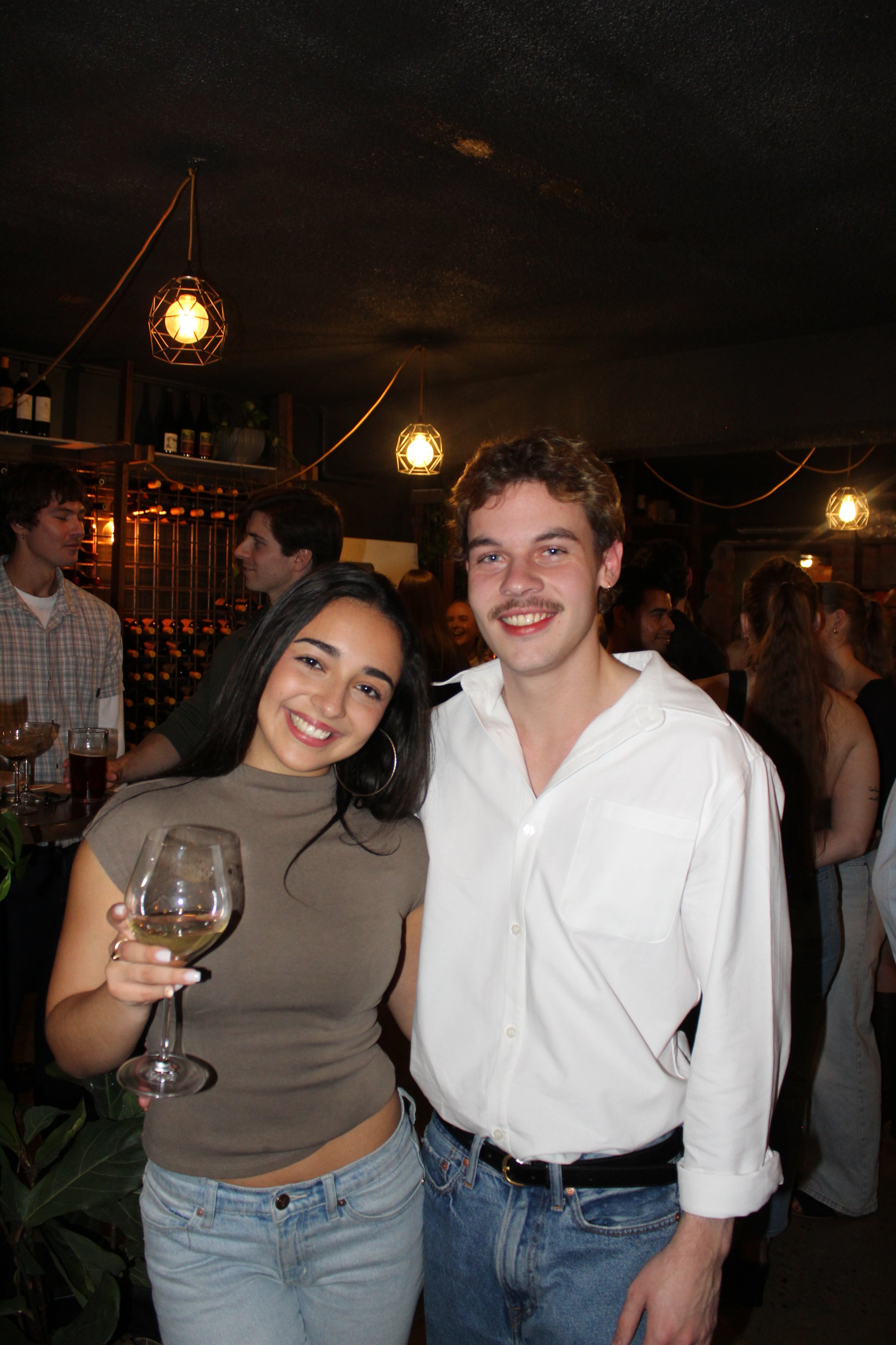 A woman holding a glass of white wine and a man smiling at a social gathering or party in a dimly lit restaurant or bar with decorative hanging pendant lights in the background.