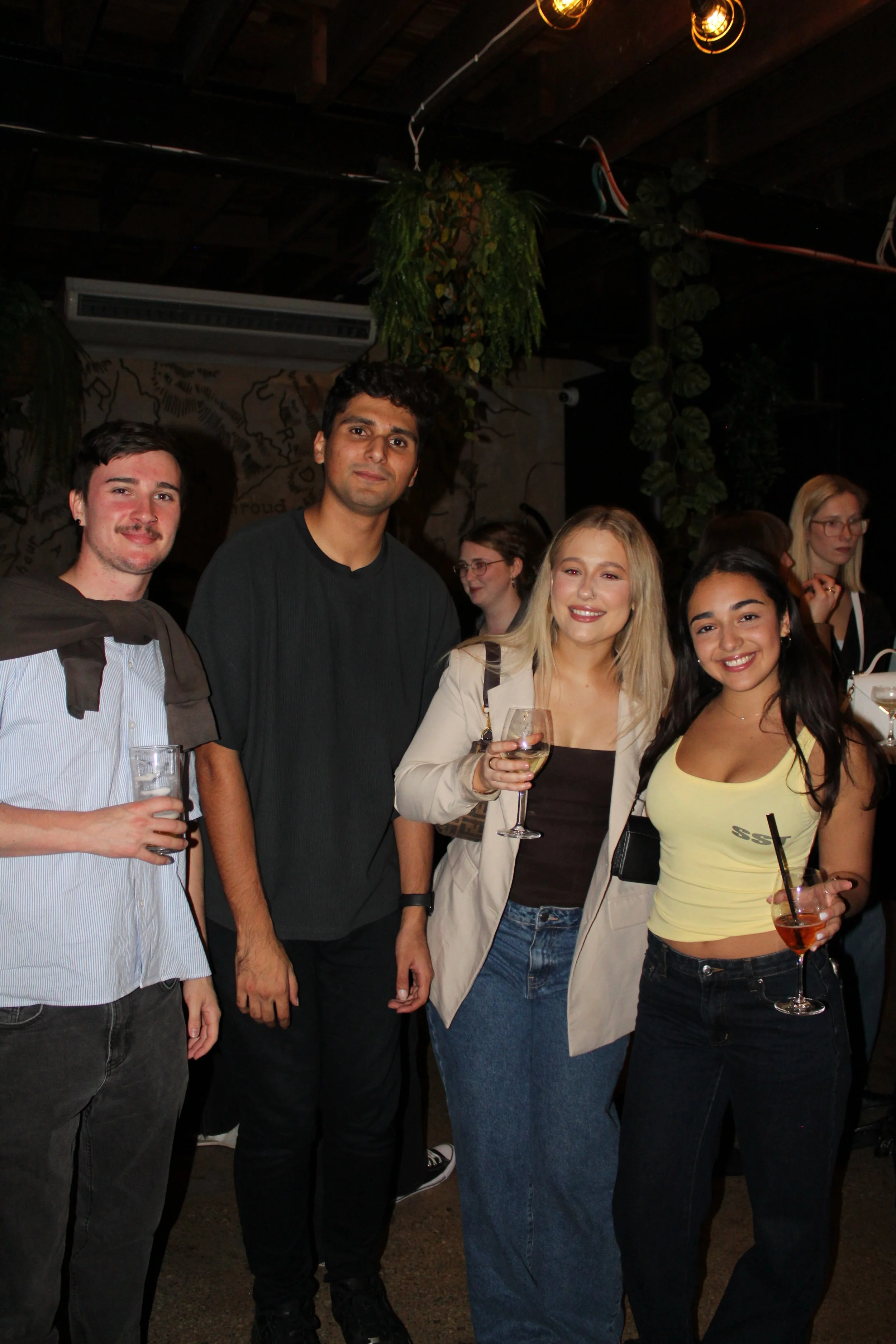 Four young adults smiling and holding drinks at a party or social gathering in a dimly lit indoor venue.