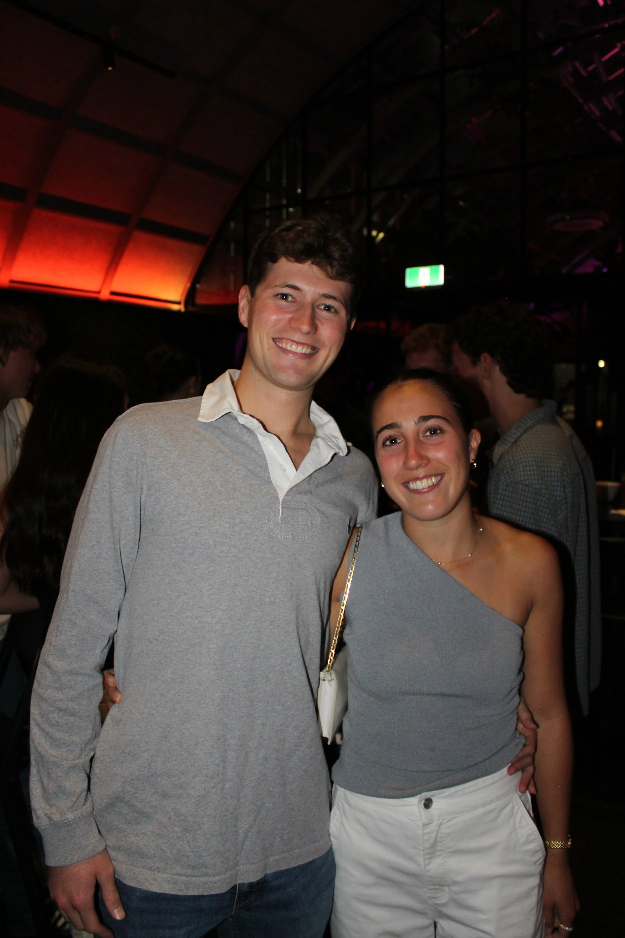 A young man and woman smiling at a social gathering or party. The man has short dark hair and is wearing a gray sweater over a white collared shirt. The woman has dark hair pulled back and is wearing a one-shoulder gray top and white pants. Other peo