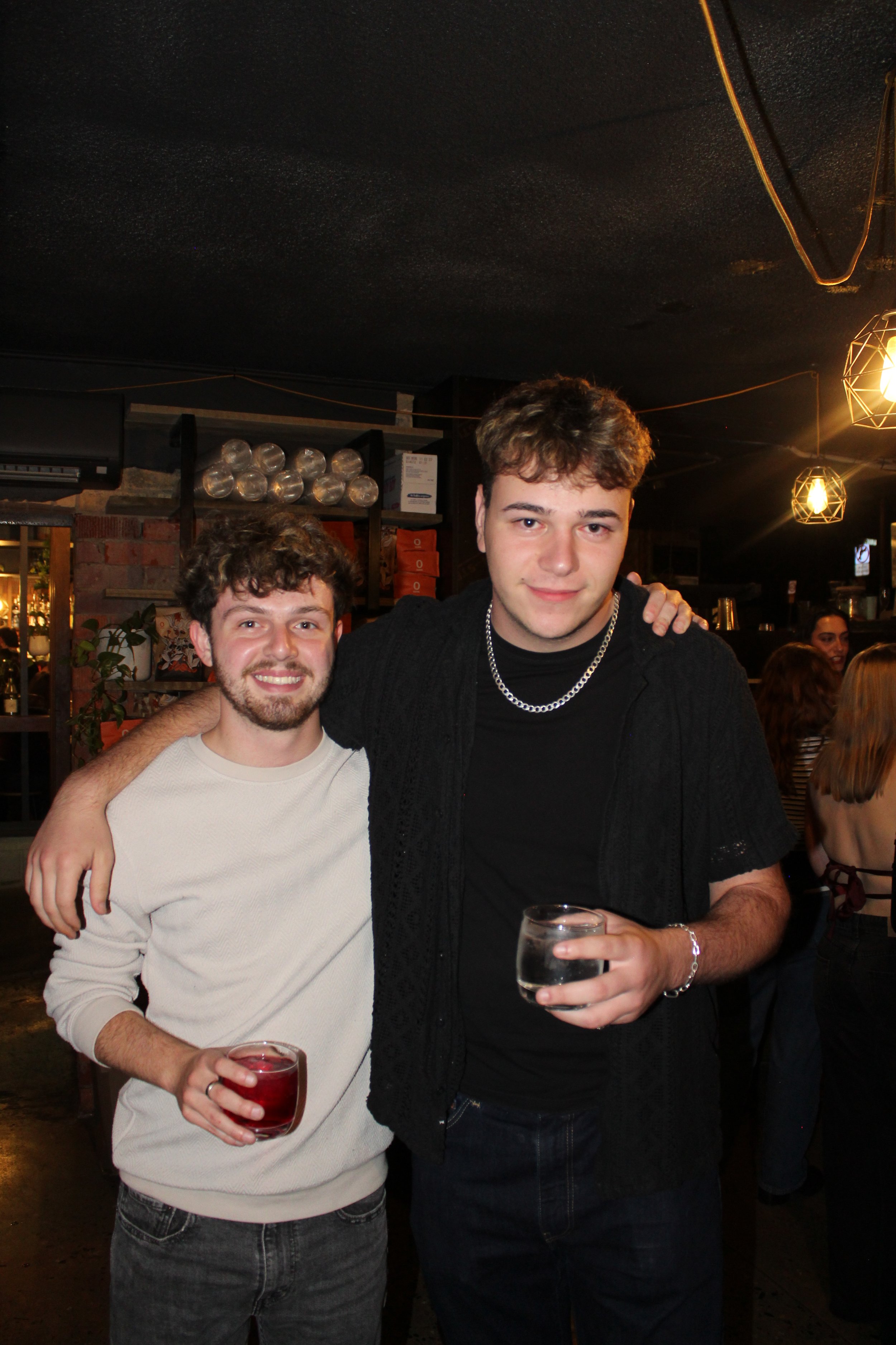 Two young men posing together at a bar or restaurant, each holding a drink, with people in the background and warm lighting.