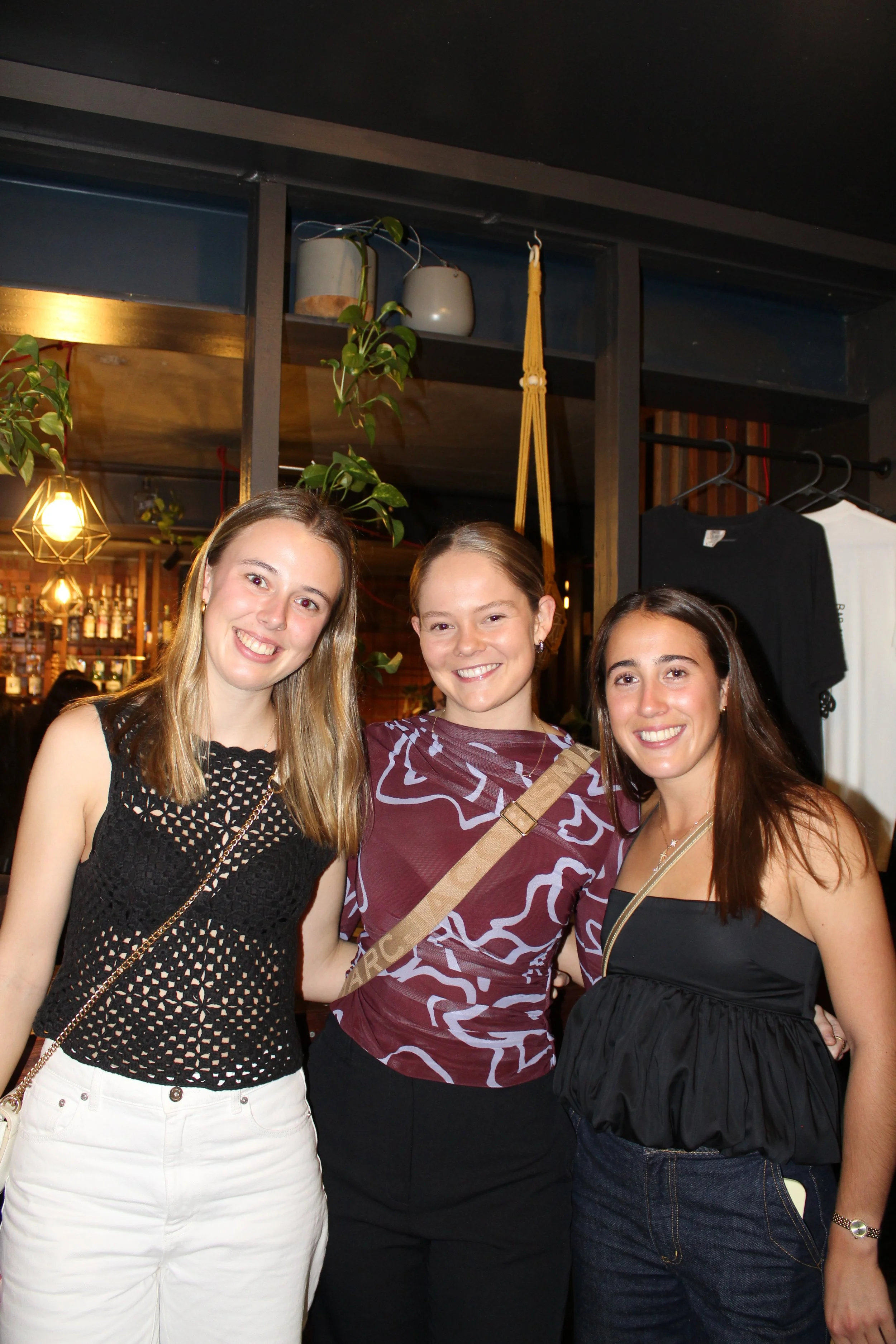 Three women smiling at the camera indoors, with a bar in the background and green plants above them.