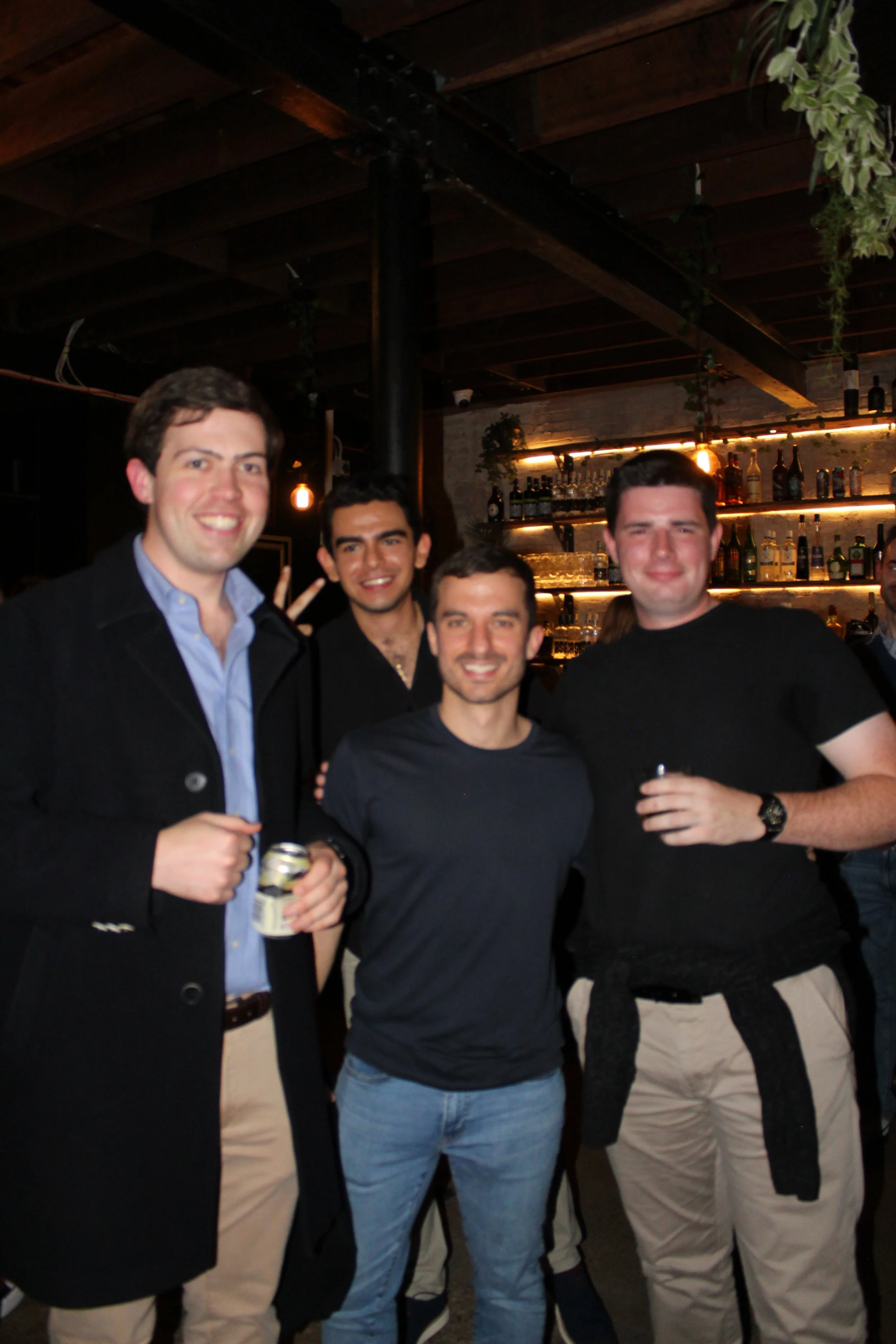 Four men standing together at a bar, smiling. Two of them are holding drinks, and a bar with bottles and glasses is in the background.