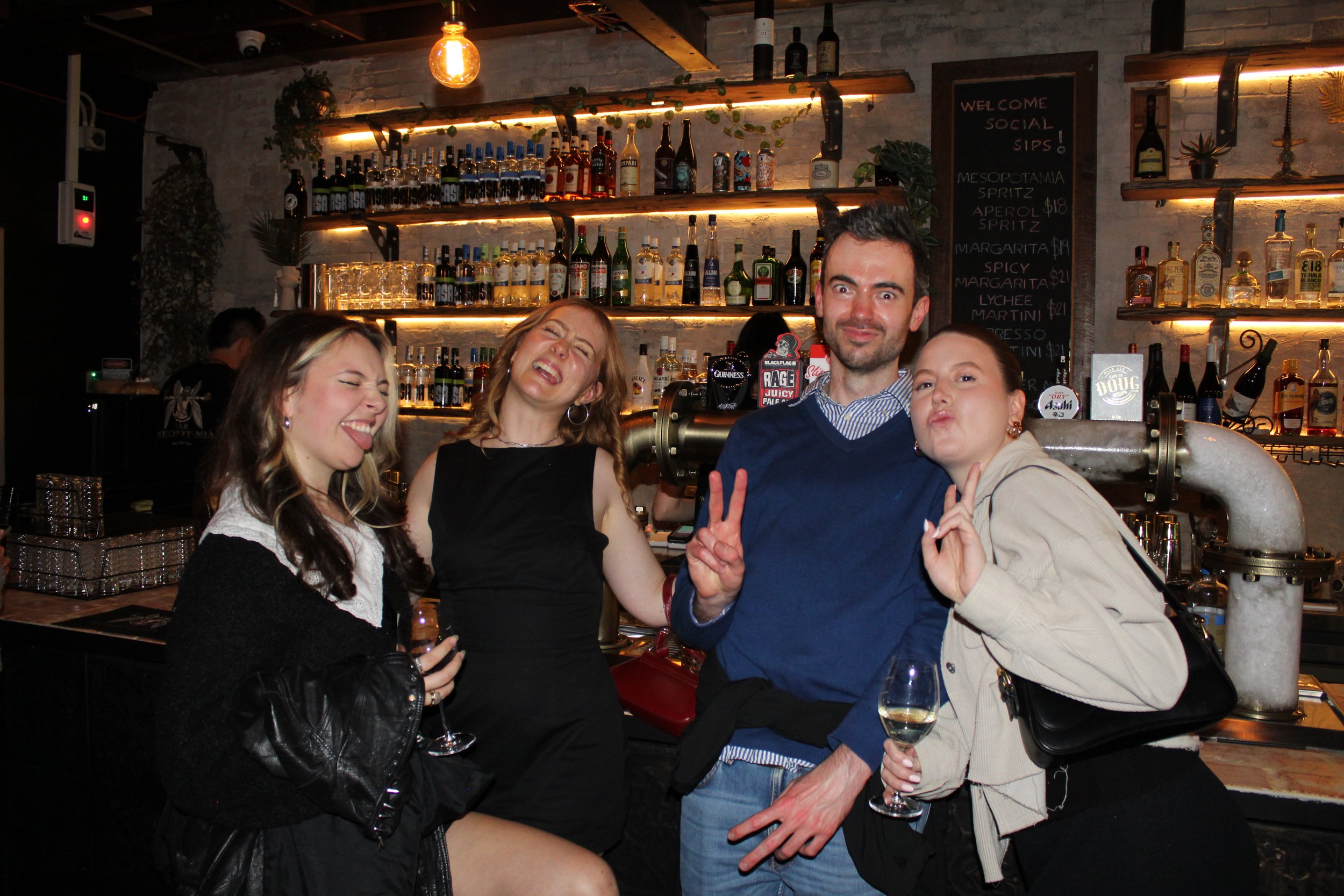 Four young adults posing and making playful facial expressions at a bar, with a decorated bar counter, bottles of alcohol, and a chalkboard menu in the background.