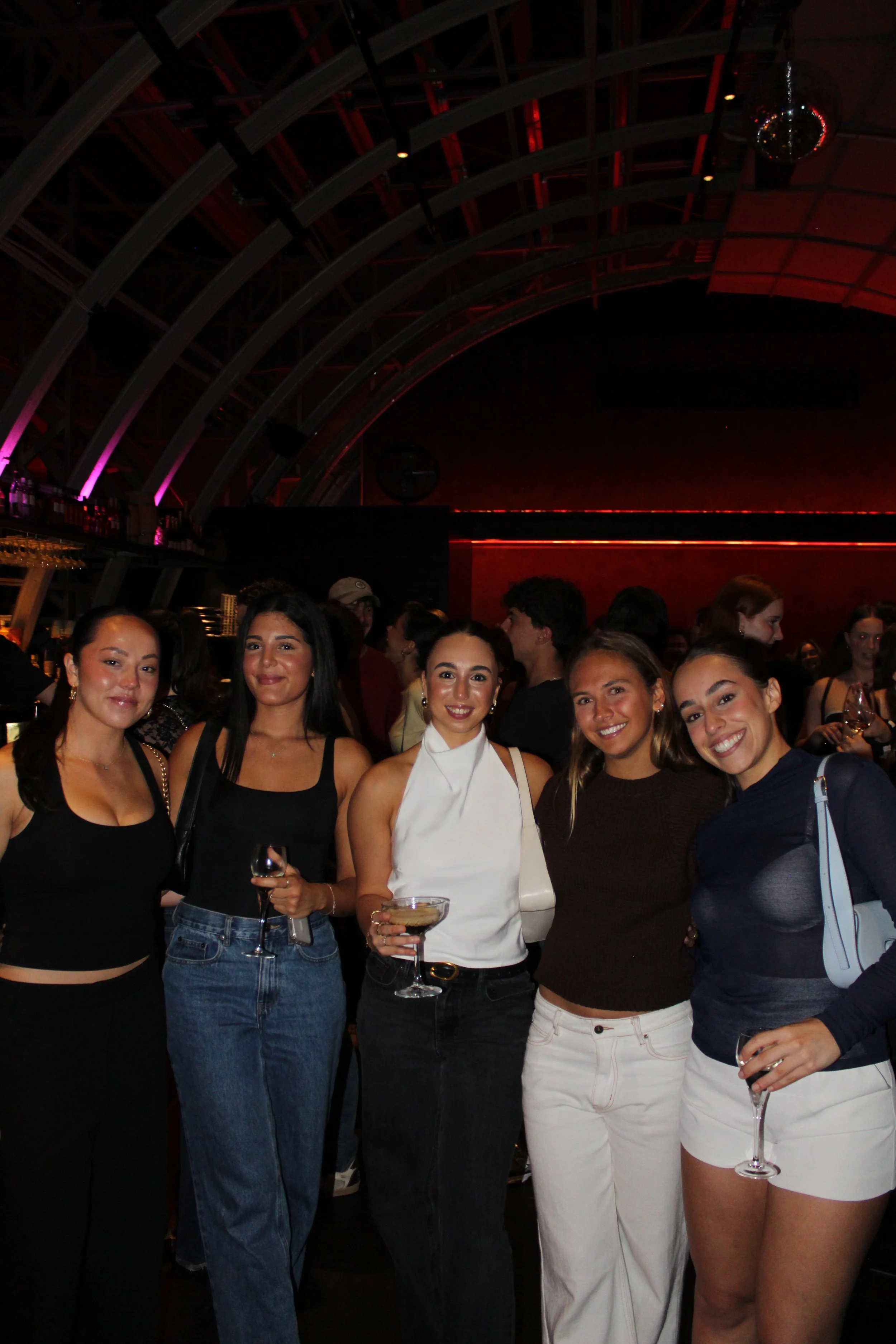 Five young women posing and smiling at a social event or party, holding drinks, in a dimly lit venue with a curved ceiling and red lighting.
