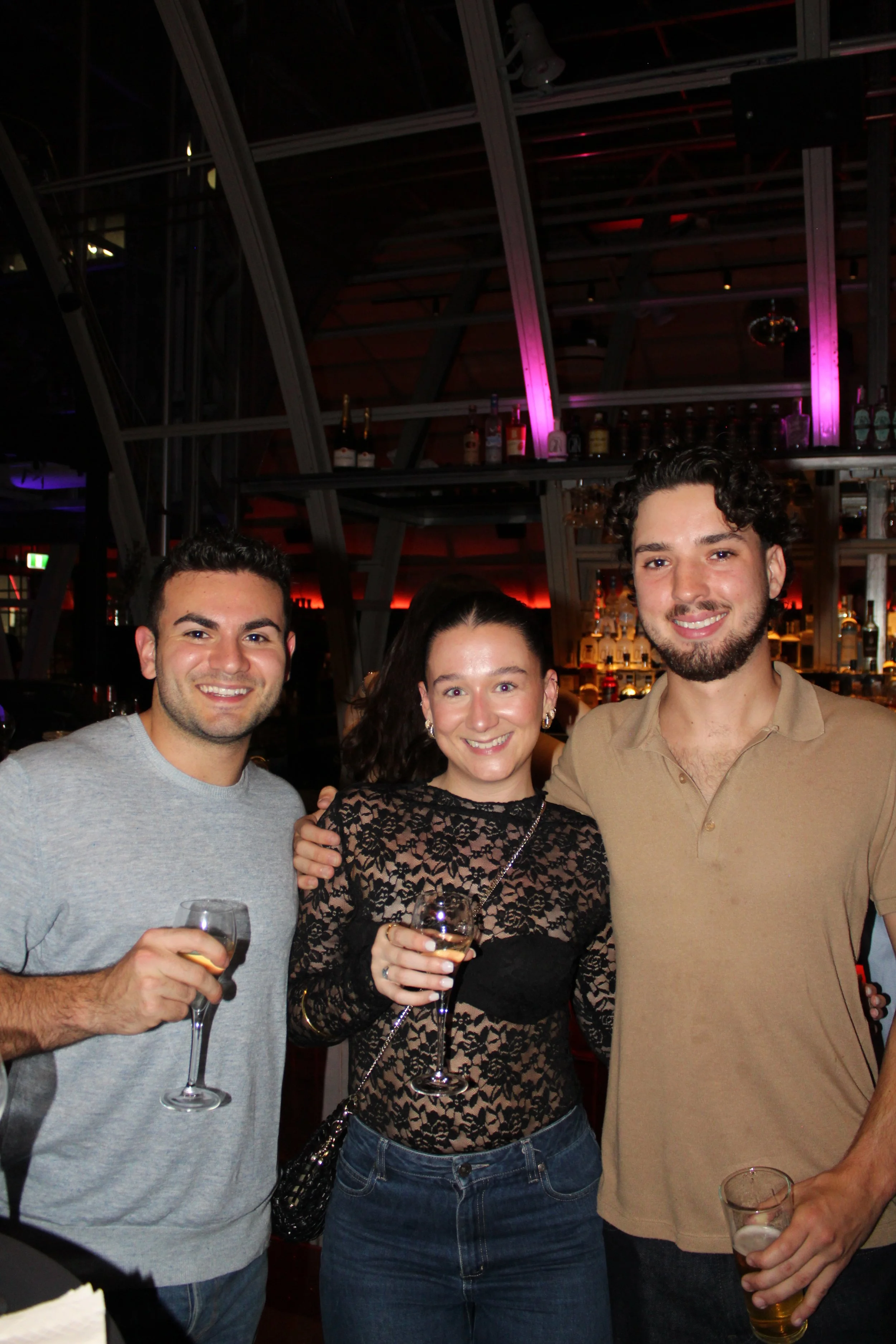 Three friends smiling at a bar, holding glasses of wine or beer, with a bar in the background.