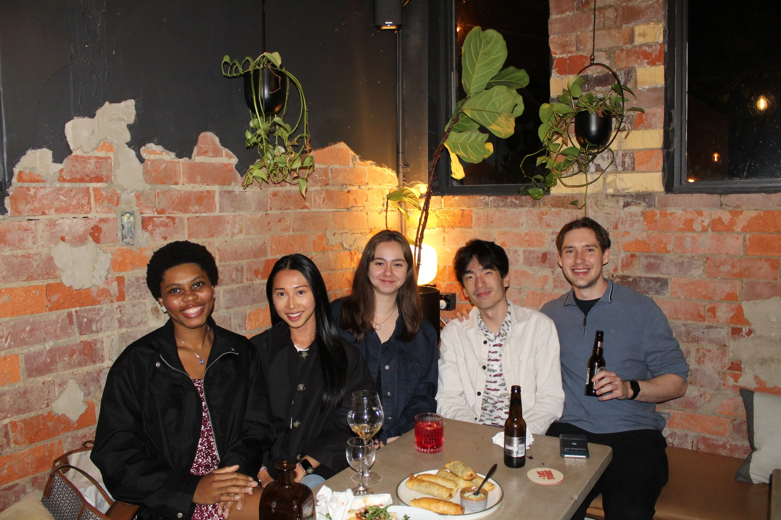 Group of five people sitting at a table with food and drinks, posing for a photo in a restaurant with a brick wall background and potted plants.