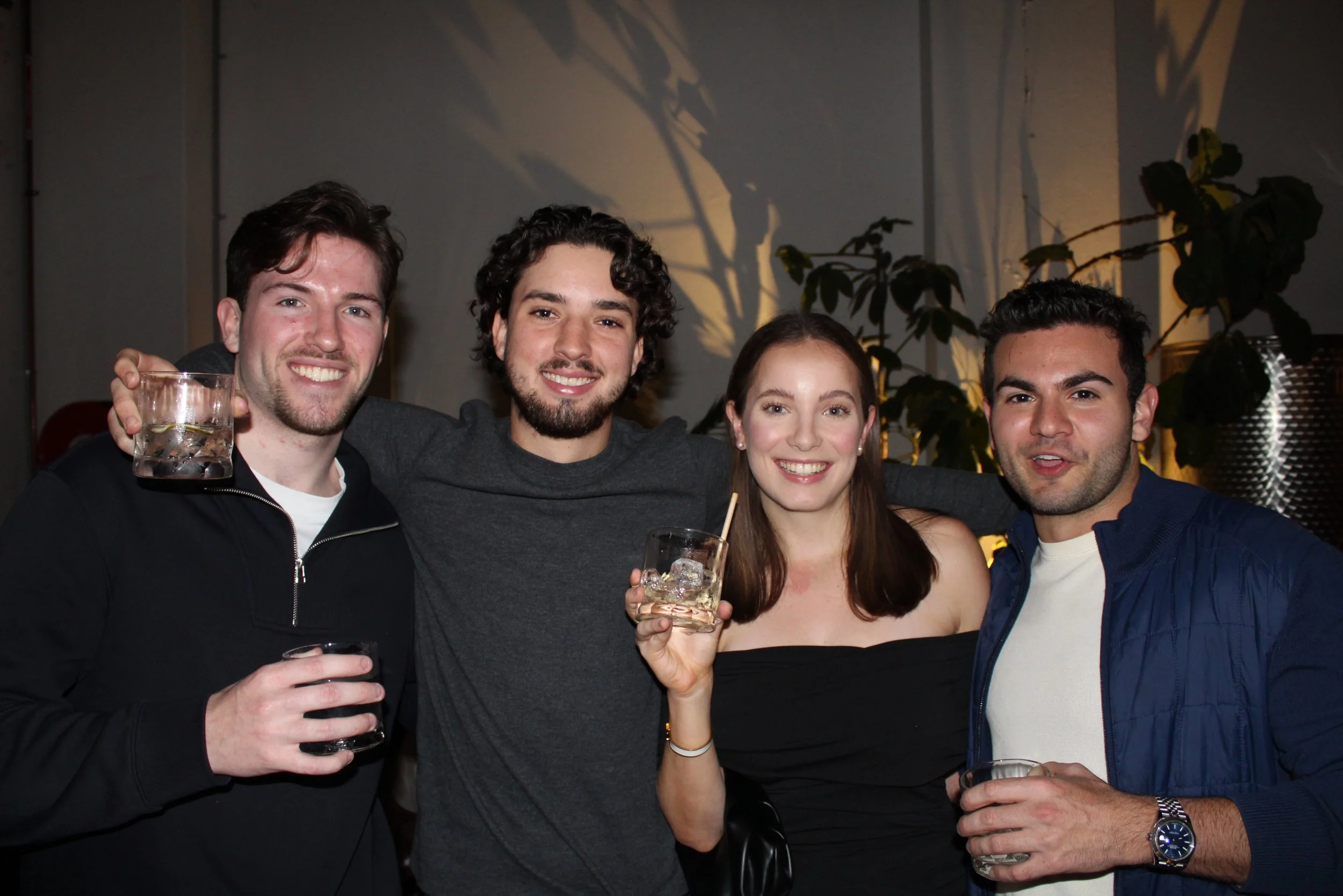 Four friends smiling at a party, holding drinks, indoors with plants and dim lighting.