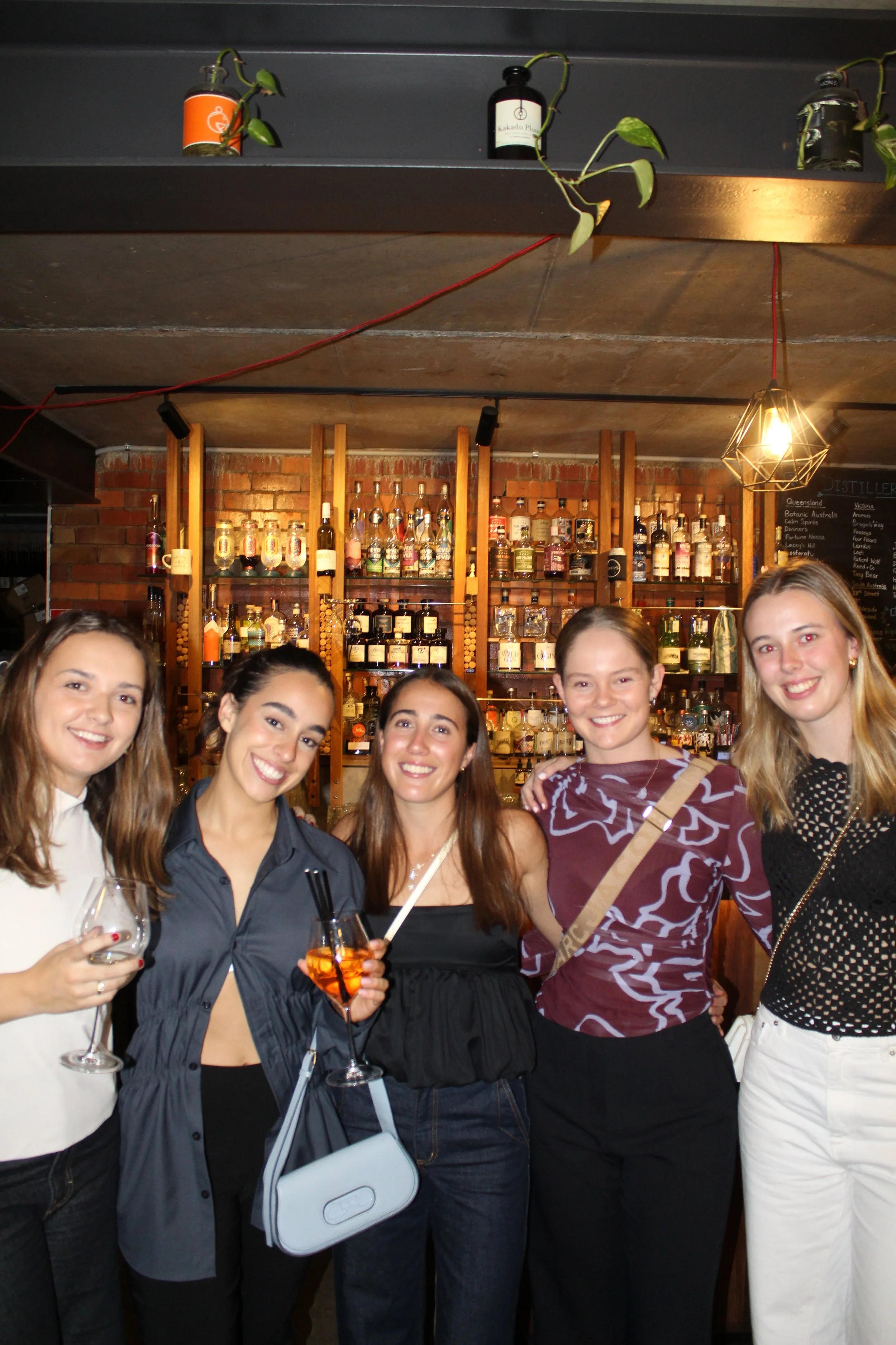 Group of five women smiling and posing together in a bar with shelves of liquor bottles behind them. They are holding drinks and dressed in casual/party attire.