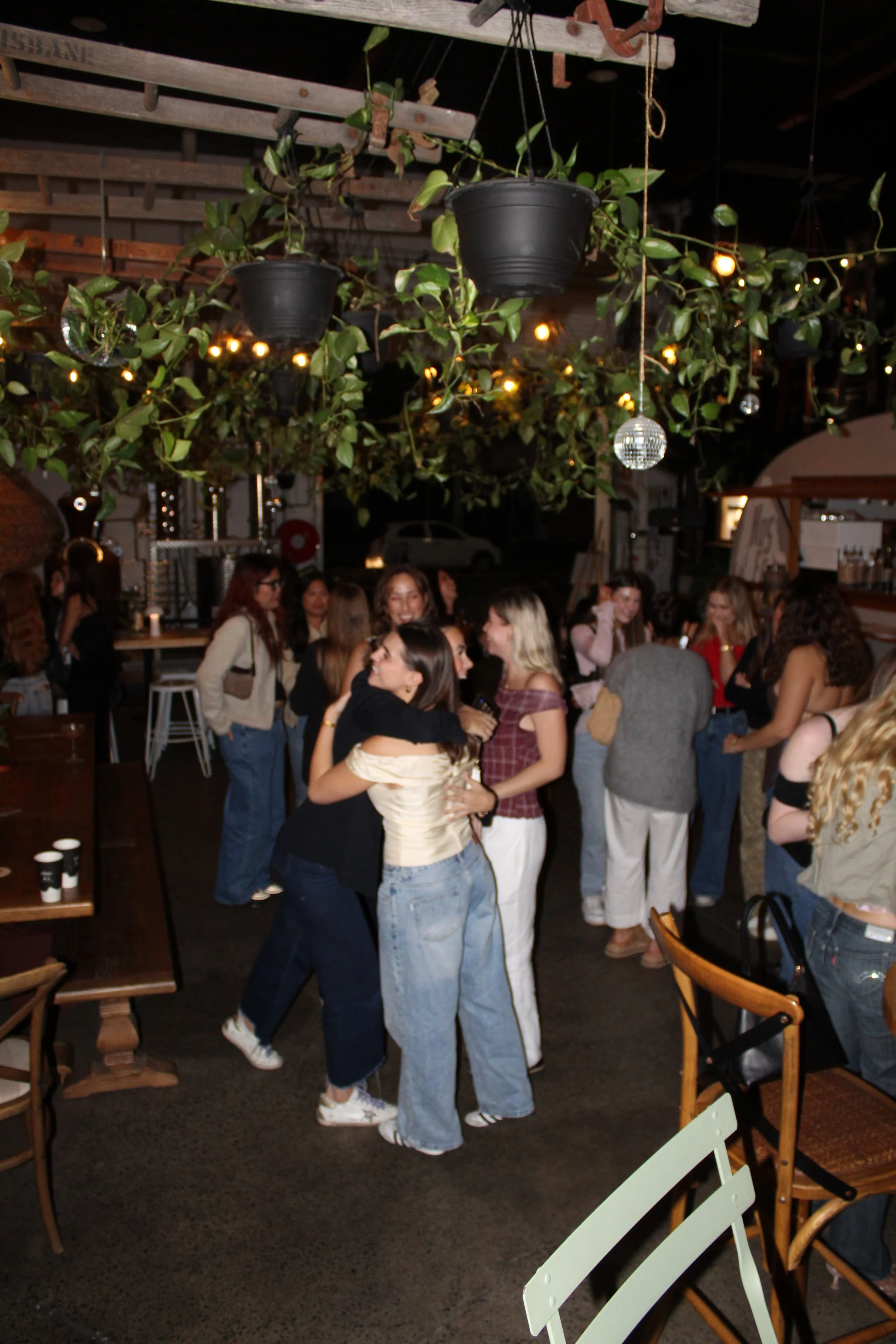 A group of women hugging and socializing inside a cozy, dimly lit bar or restaurant with hanging plants, string lights, and disco balls.