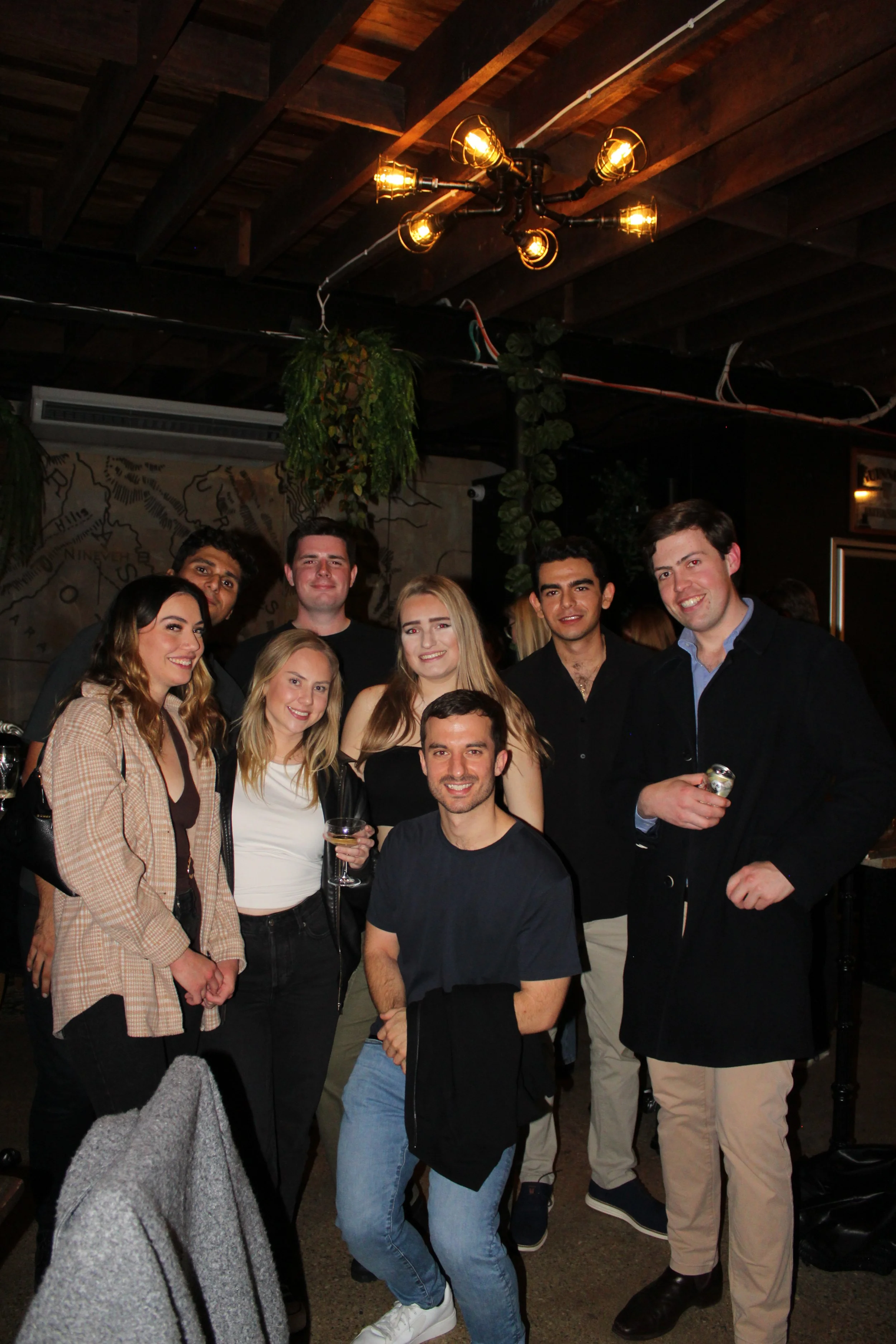 A group of nine friends in a bar or restaurant taking a group photo. They are smiling and some are holding drinks, with a rustic wooden ceiling and greenery hanging from pipes in the background.