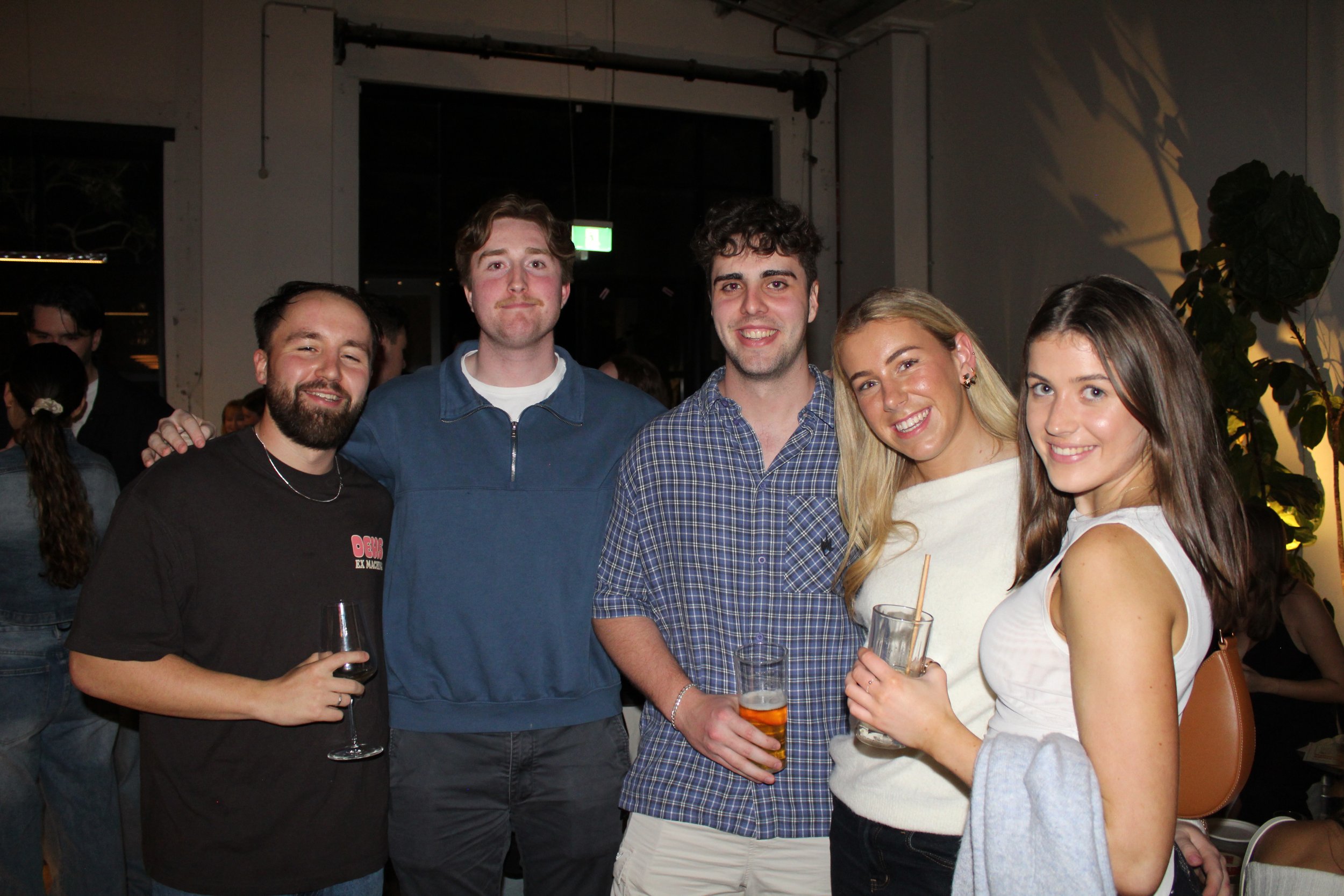 Group of five young adults smiling and enjoying drinks at a social gathering indoors.