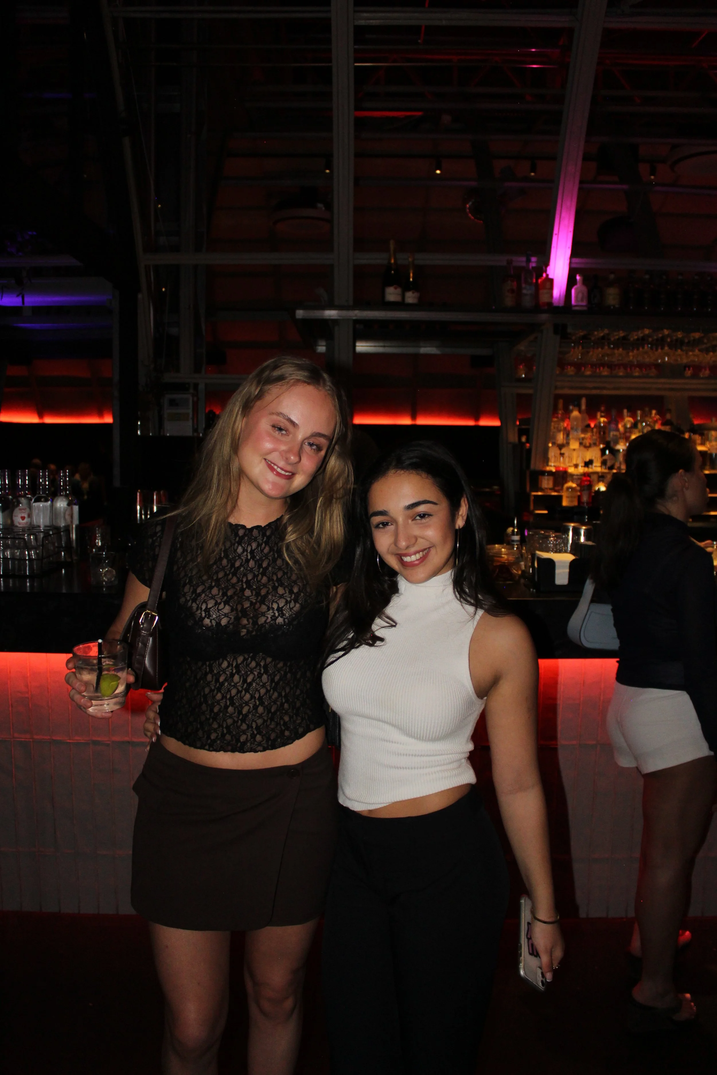 Two young women smiling at a bar during the night, one holding a drink with a lime, with drinks and bar supplies visible in the background.
