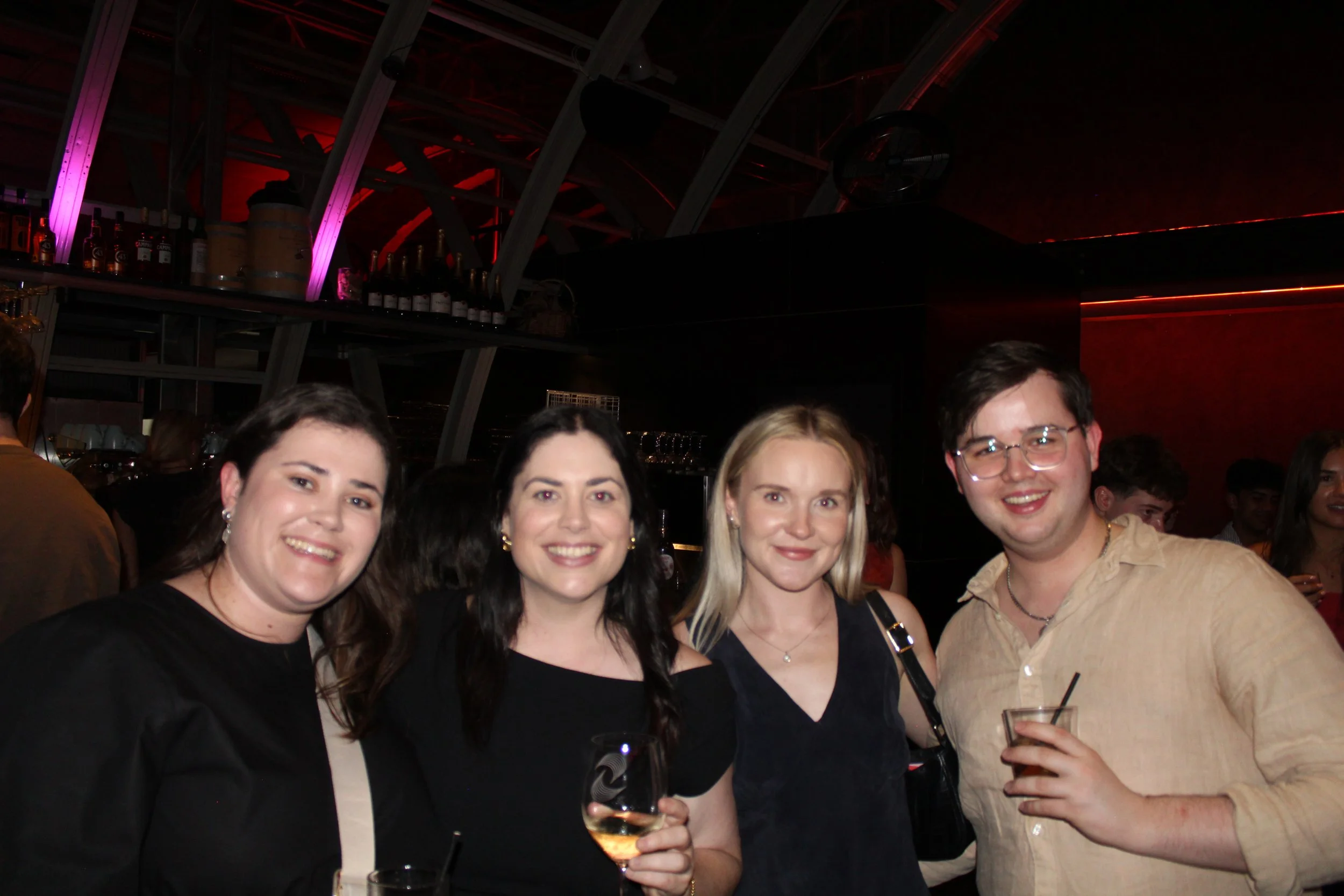 Four friends smiling and posing for a photo at a bar or nightclub, with drinks in hand, under colorful lighting.
