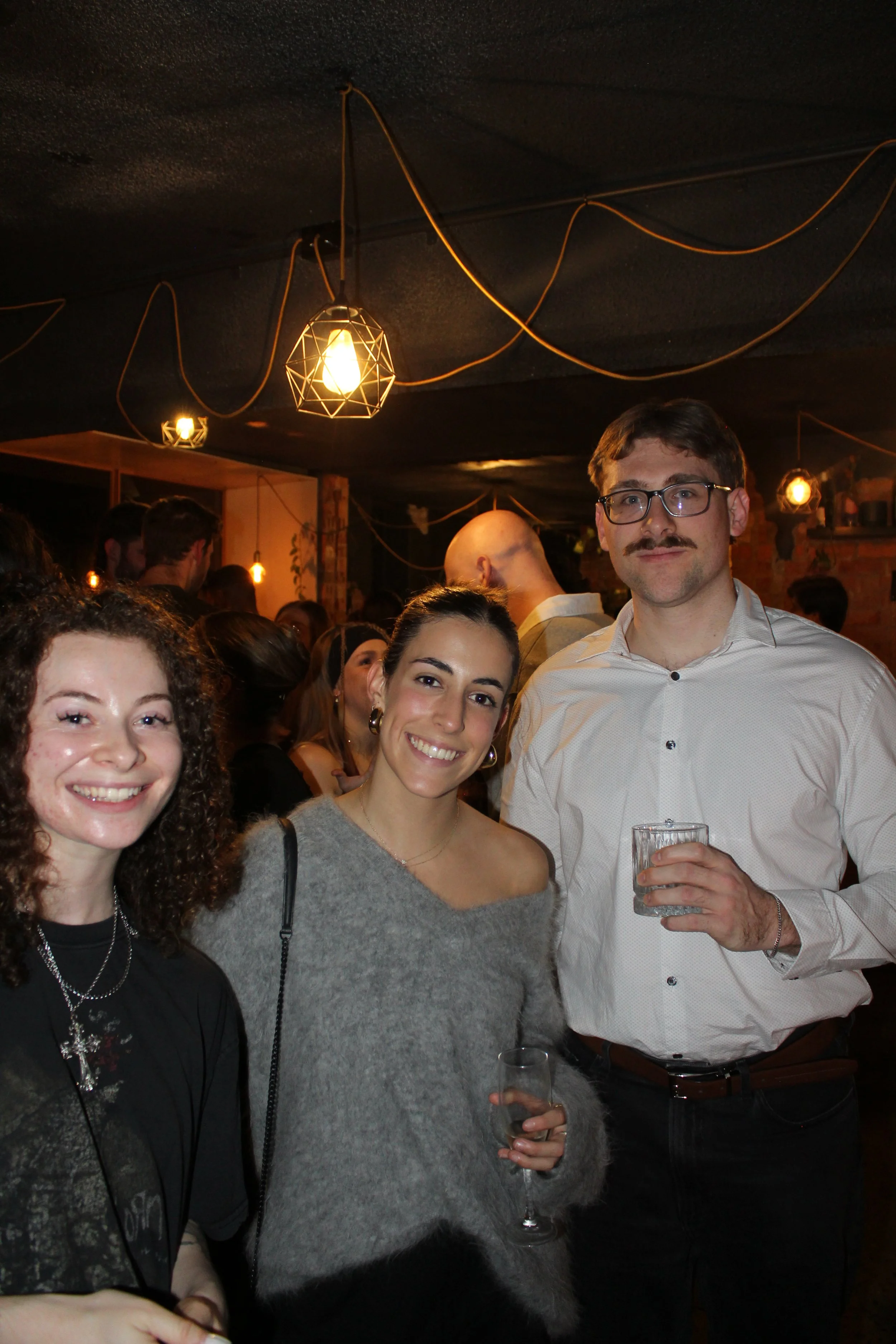 People attending a social gathering or party in a dimly lit indoor space with hanging light fixtures and exposed brick walls.