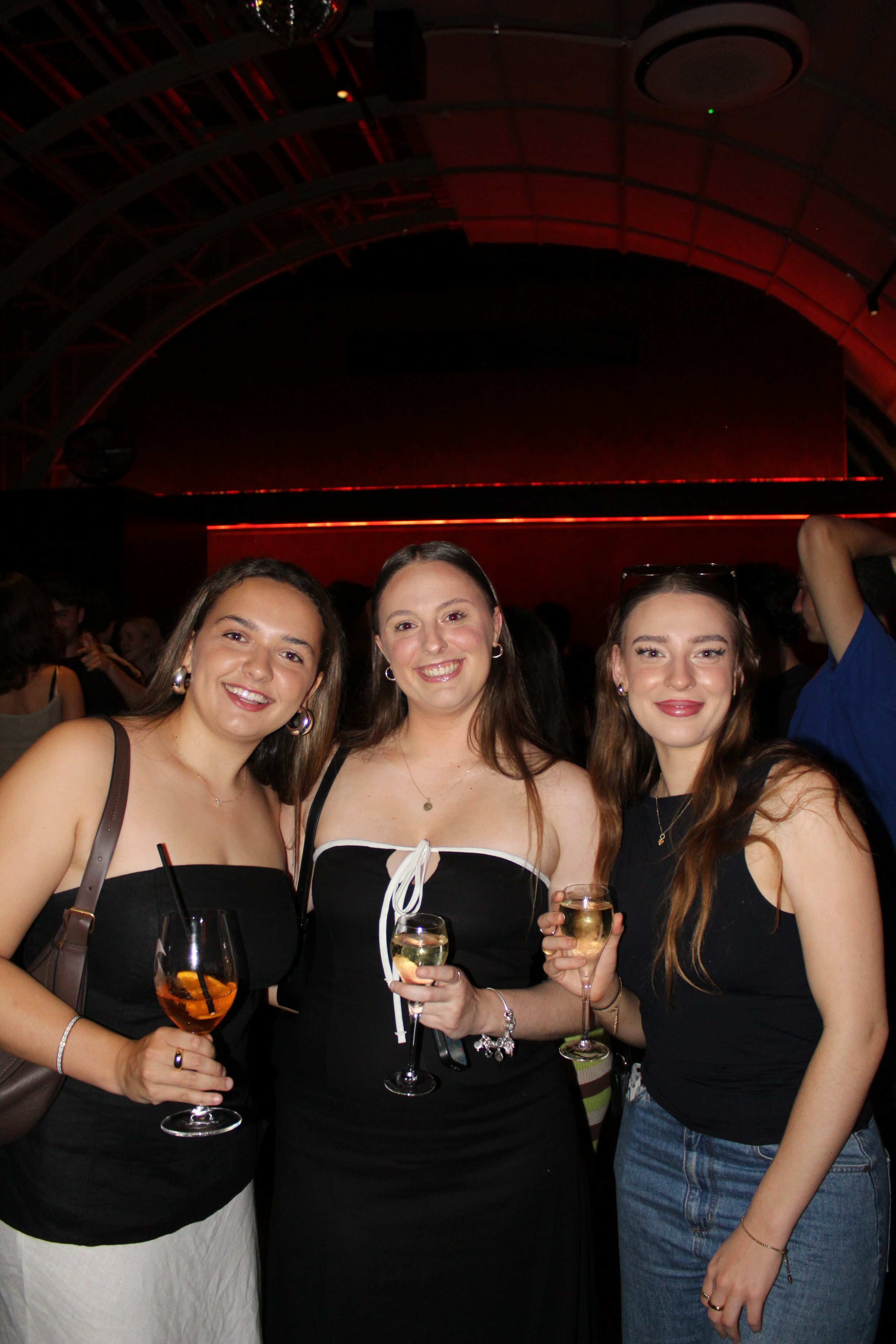 Three women smiling and holding drinks at a crowded party or bar with red lighting in the background.