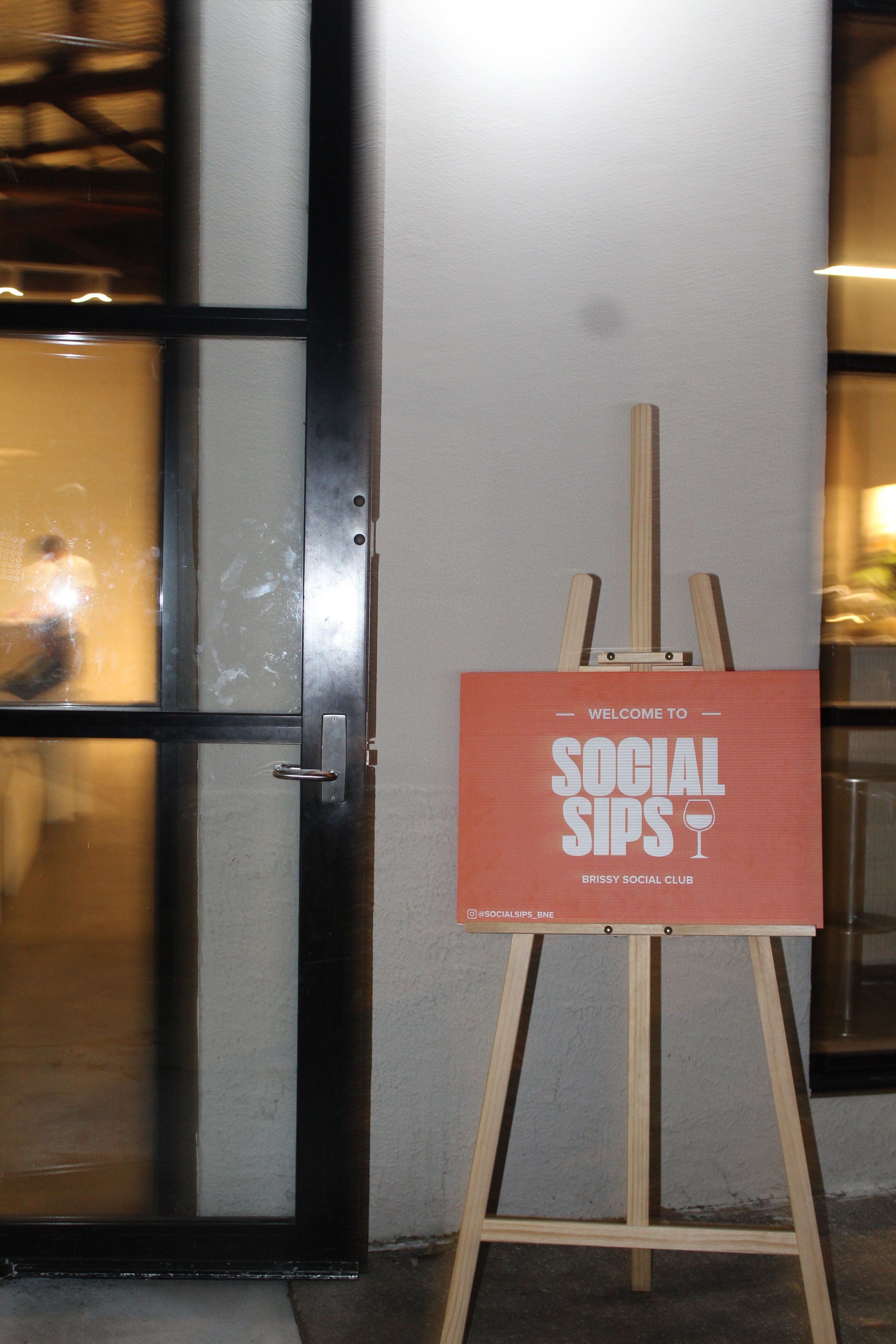 Easel with a sign reading 'Welcome to Social Sips' outside a glass door.