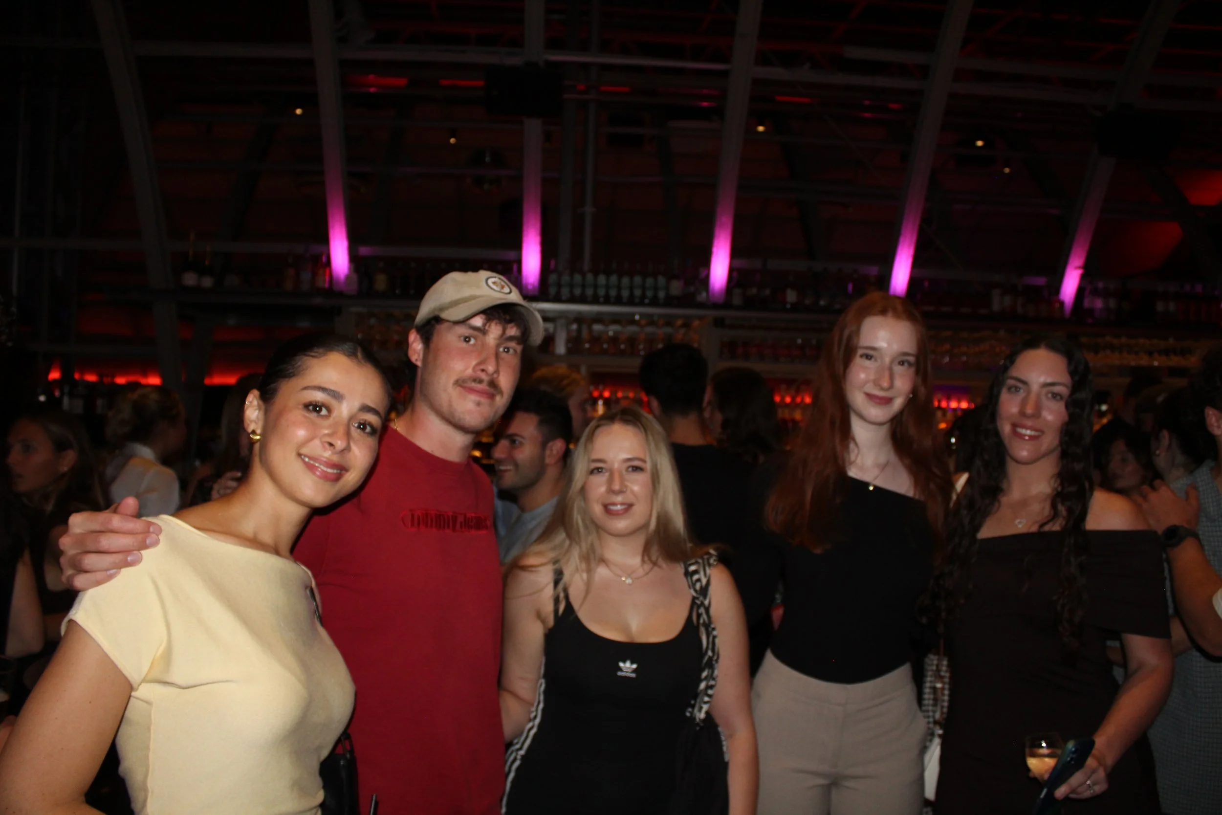 Group of five young adults, three women and two men, at a crowded indoor social event with purple and red lighting.