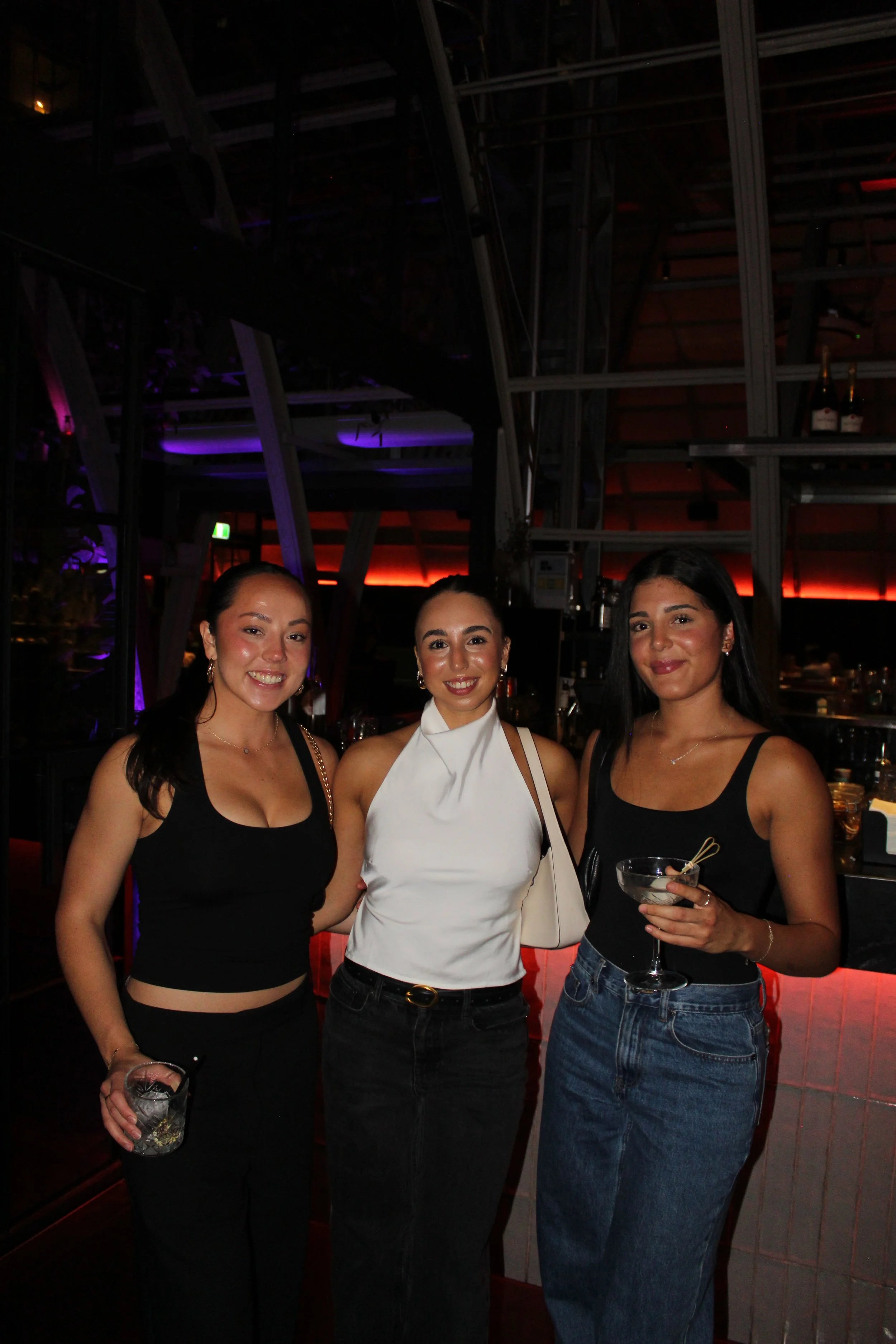 Three women standing together in a bar, smiling at the camera, holding drinks, with a dark, modern interior and dim lighting.