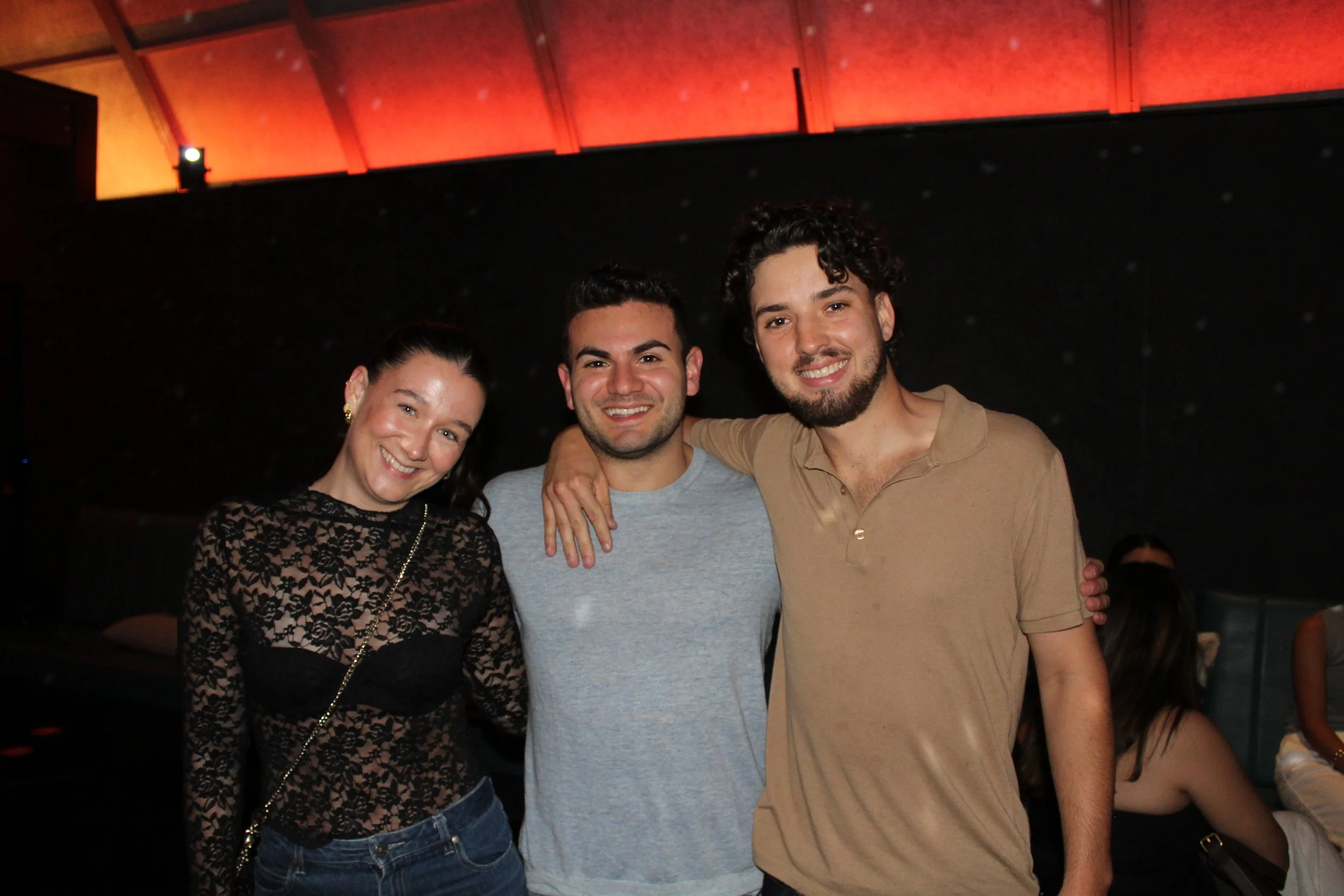 Three young adults smiling and posing together indoors, with two men in the middle and a woman on the left. The woman is wearing a black lace top, the man in the middle is wearing a gray shirt, and the man on the right is wearing a beige polo shirt. 