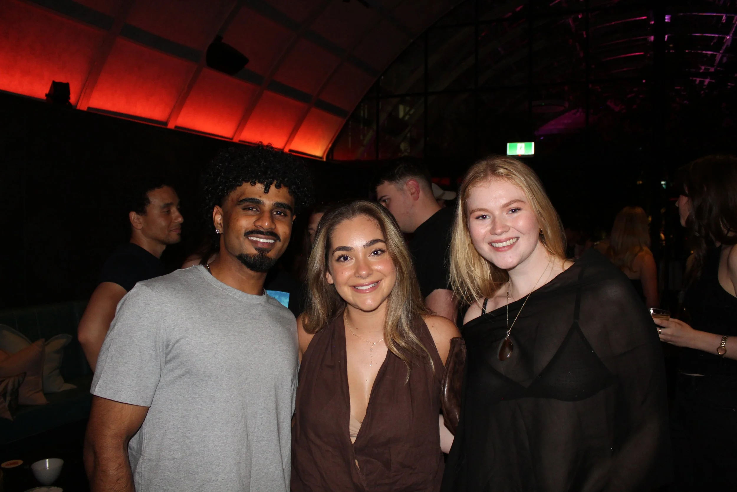 Three people smiling at a party or social gathering, two women and one man, standing close together in a dimly lit venue with a red and purple lit ceiling.