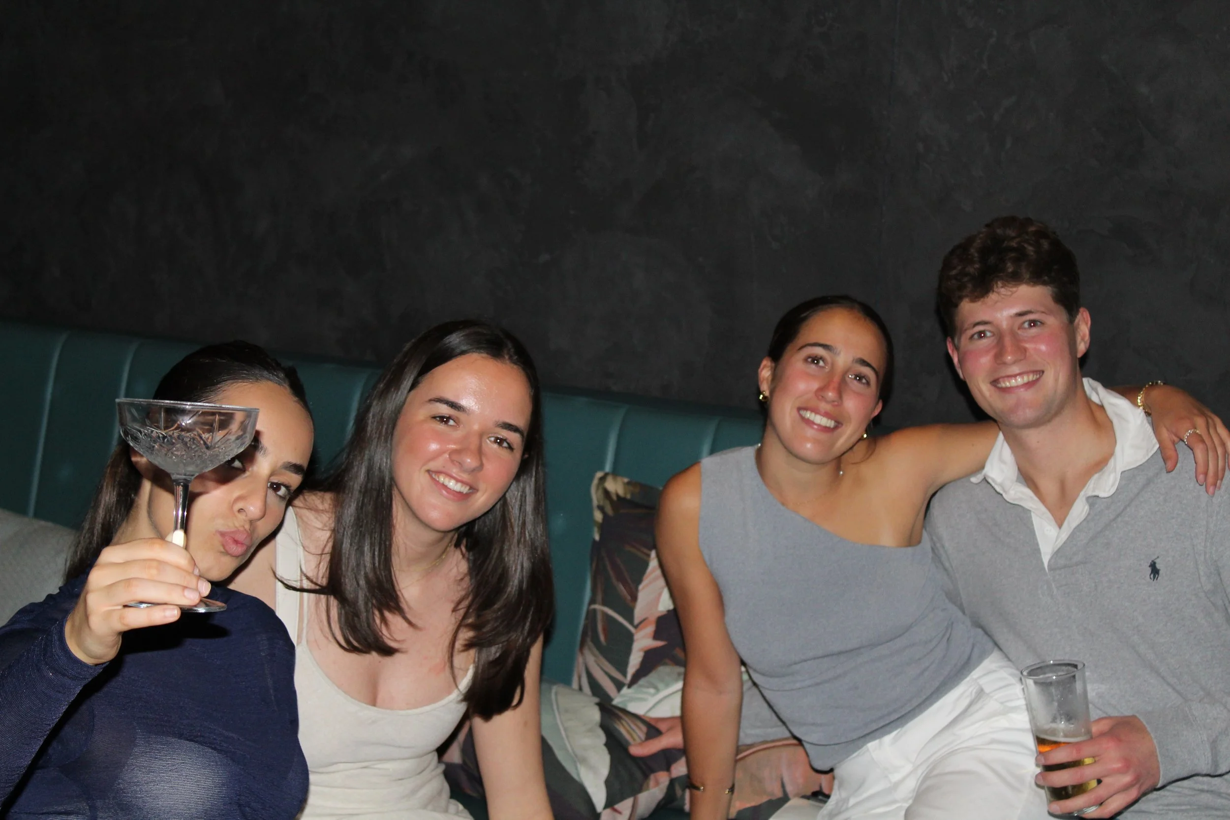 Four young adults sitting on a couch, smiling, with two women holding drinks, including a cocktail glass and a beer glass, in a dark, indoor setting.