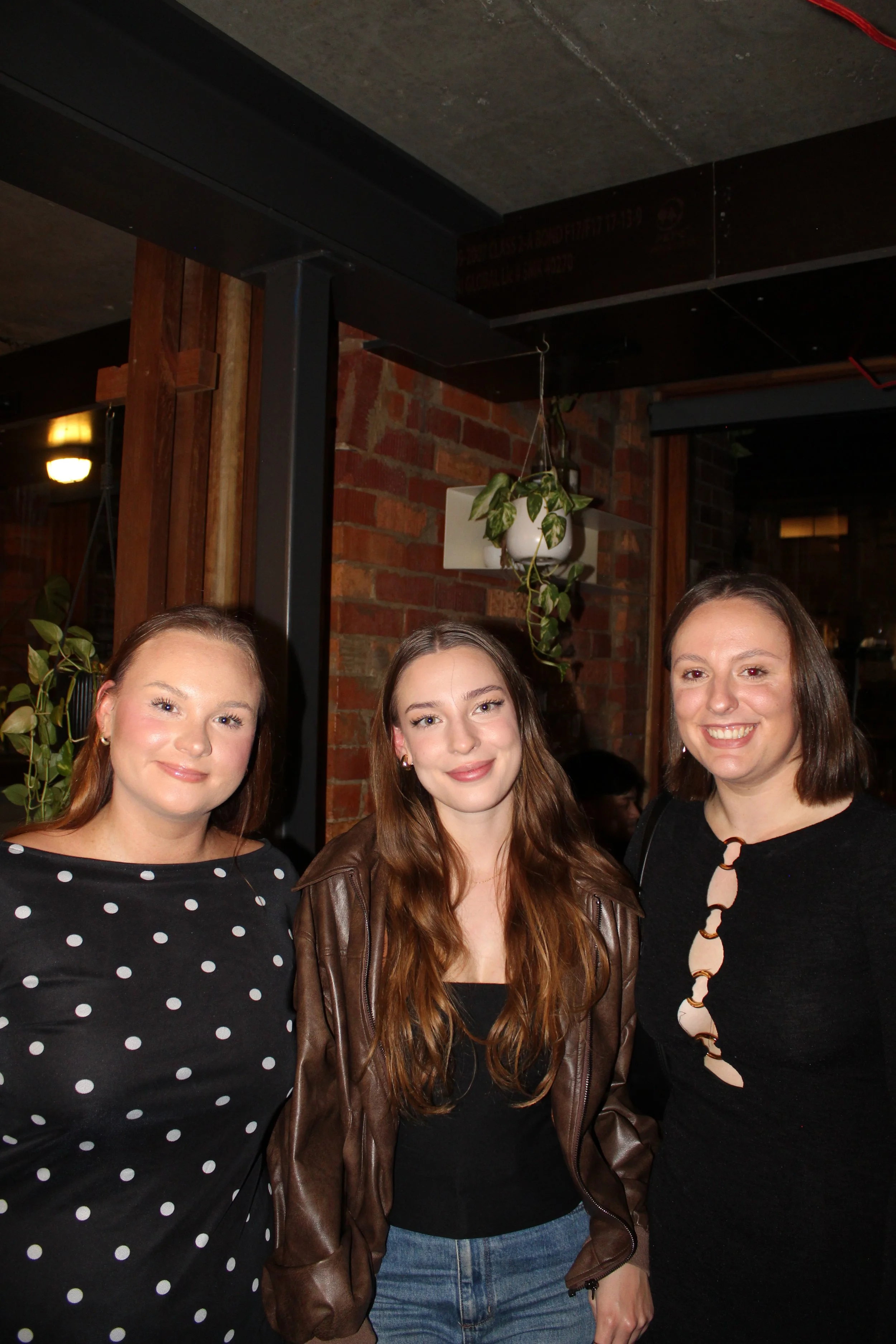 Three women smiling in a restaurant with brick walls and indoor plants.