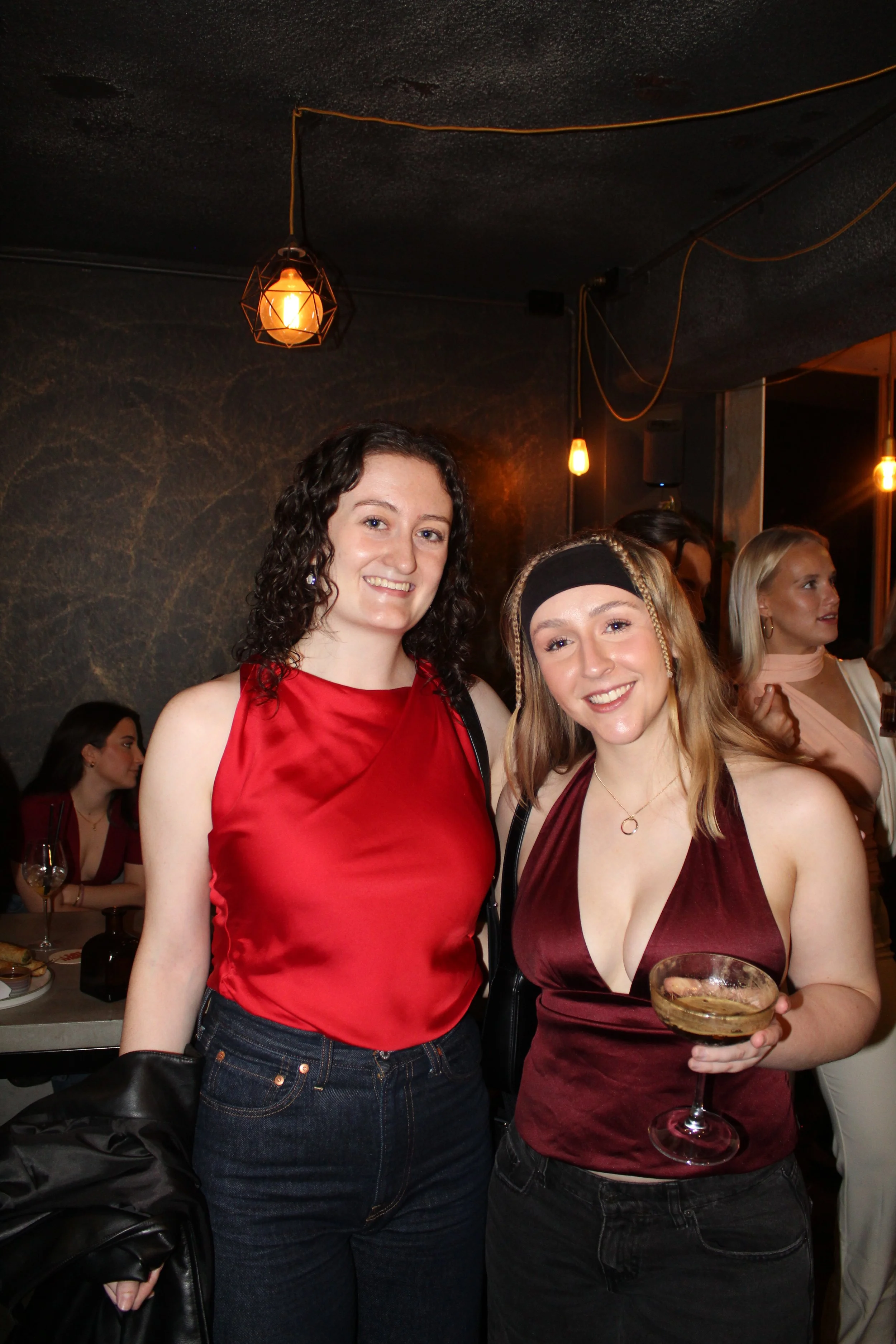 Two women smiling at a party, one in a red satin top and the other in a burgundy dress holding a champagne glass, with other guests in the background and warm lighting.