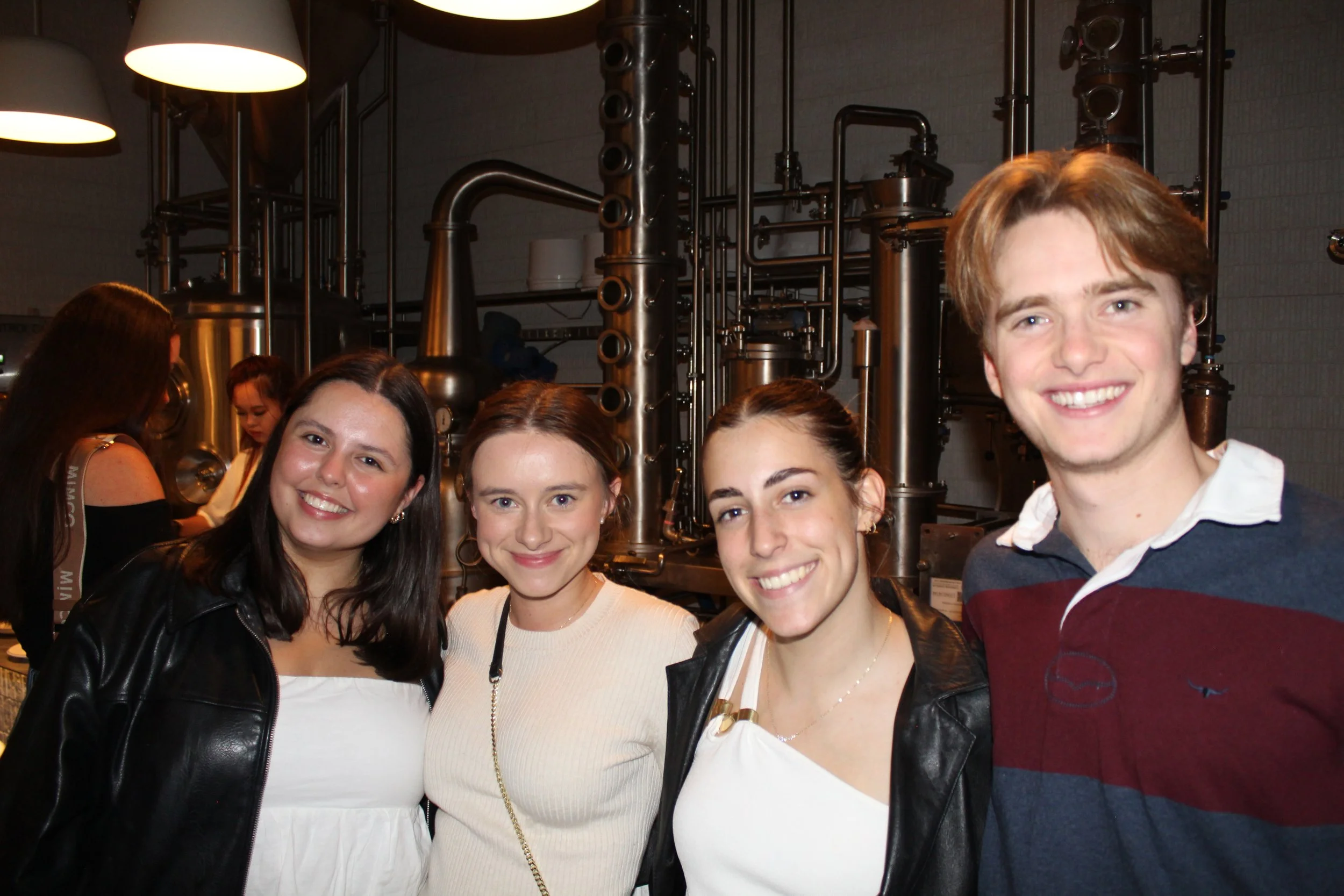 Four young adults smiling in front of a metallic industrial kitchen setup.