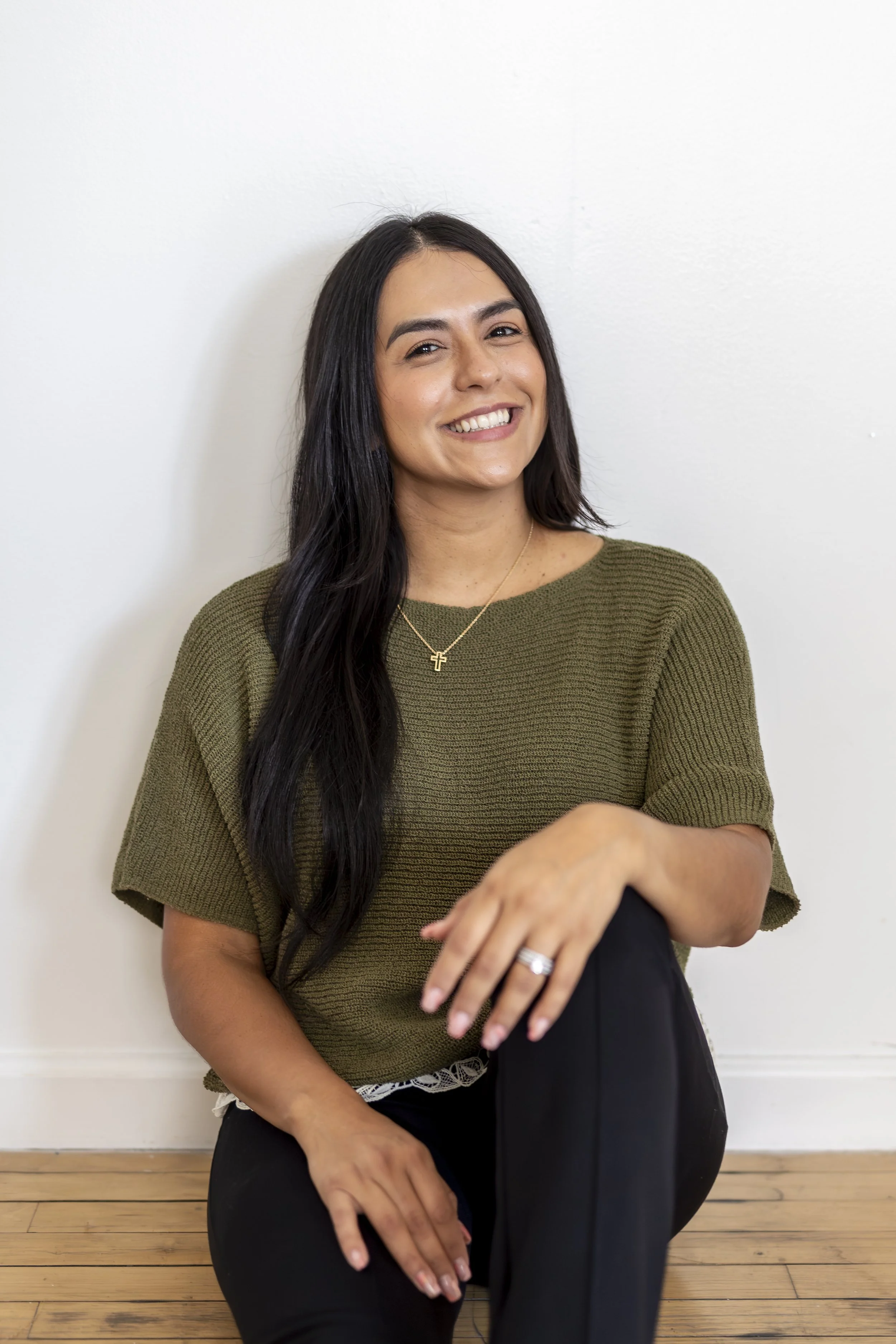A latina therapist with long black hair, smiling, wearing an olive green sweater and black pants, sitting against a white wall on a wooden floor.
