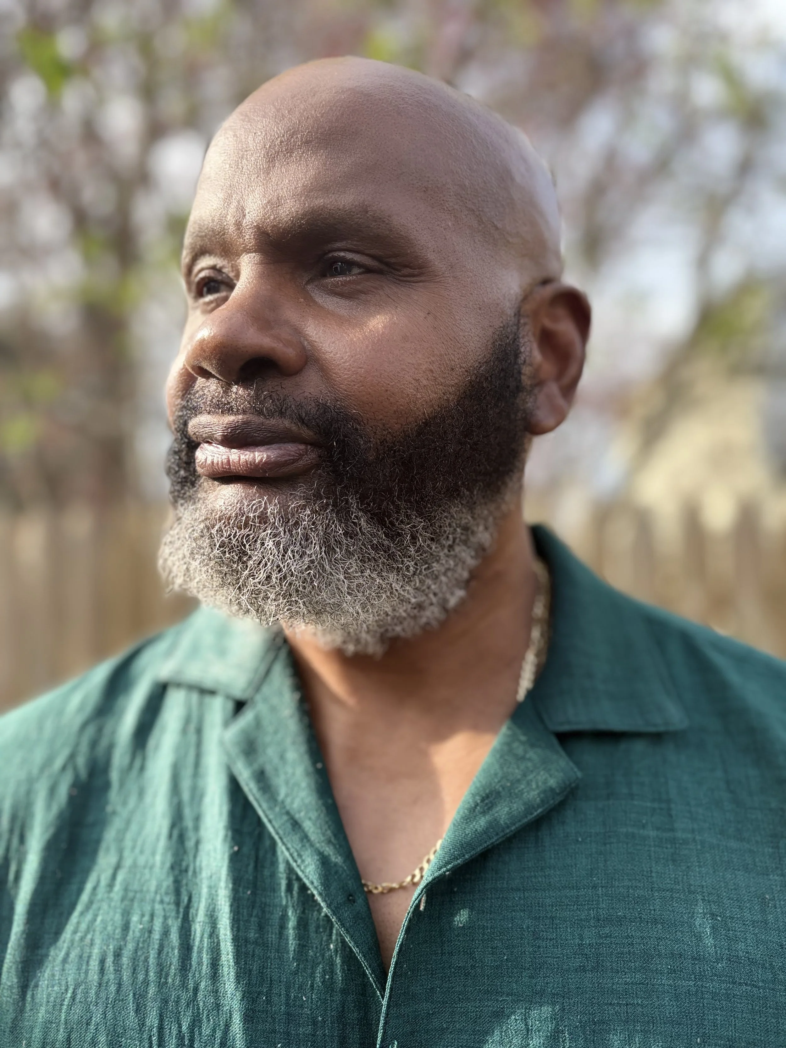 Close-up of an African American man with a beard, wearing a green collared shirt and a gold chain, outdoors during daytime.