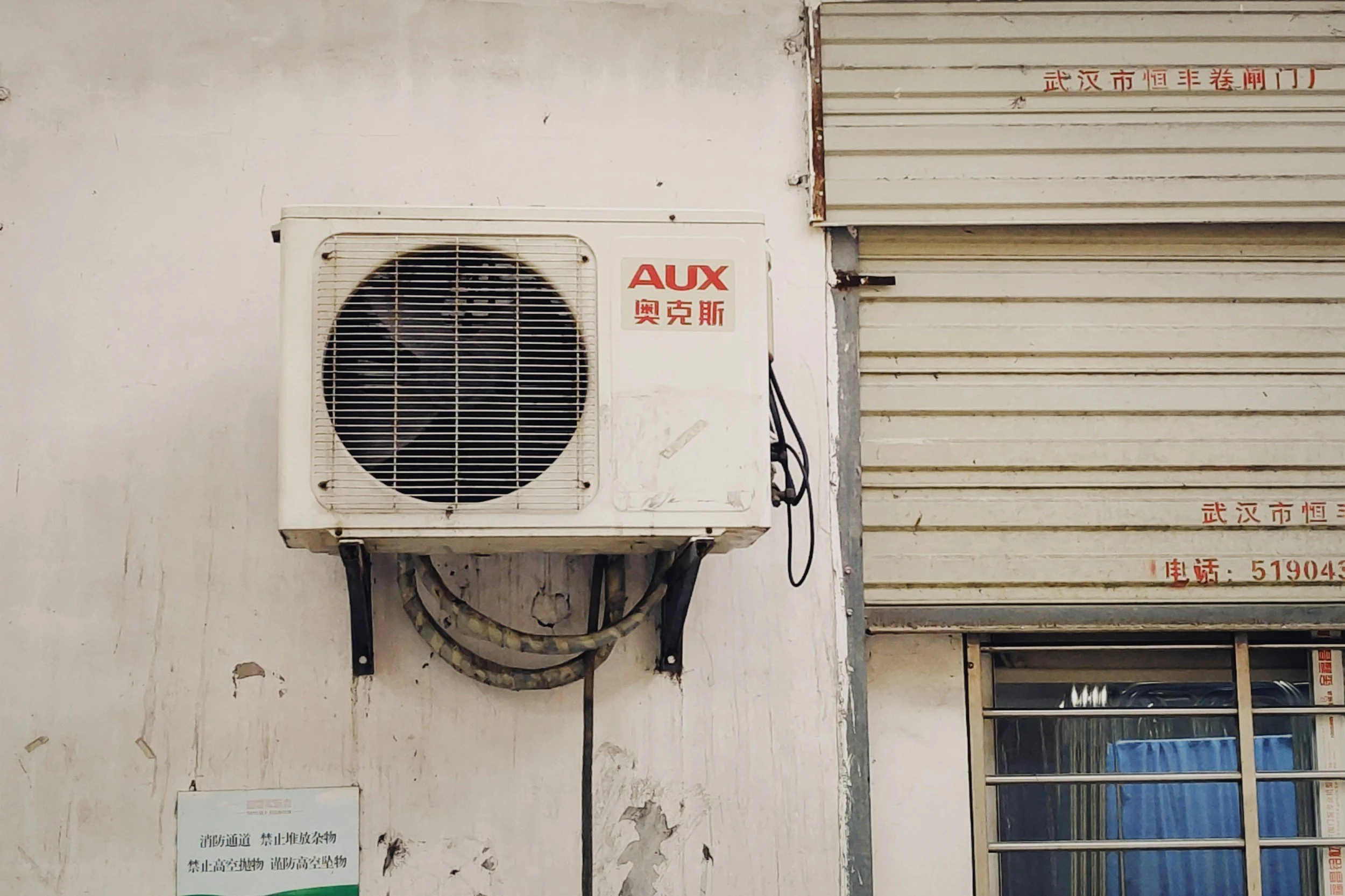 An air conditioning unit mounted on a white wall next to a metal roller shutter door with red Chinese characters.