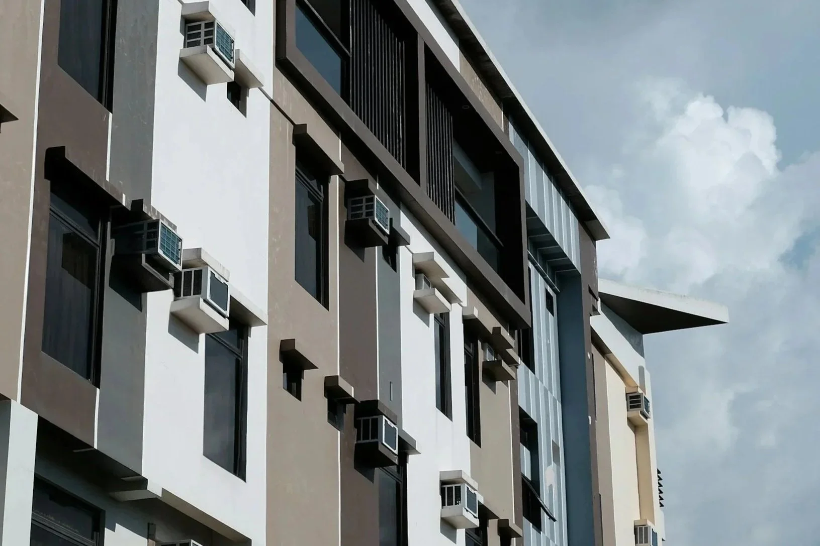View of a modern apartment building with multiple windows and air conditioning units installed on the exterior walls, against a background of a partly cloudy sky.