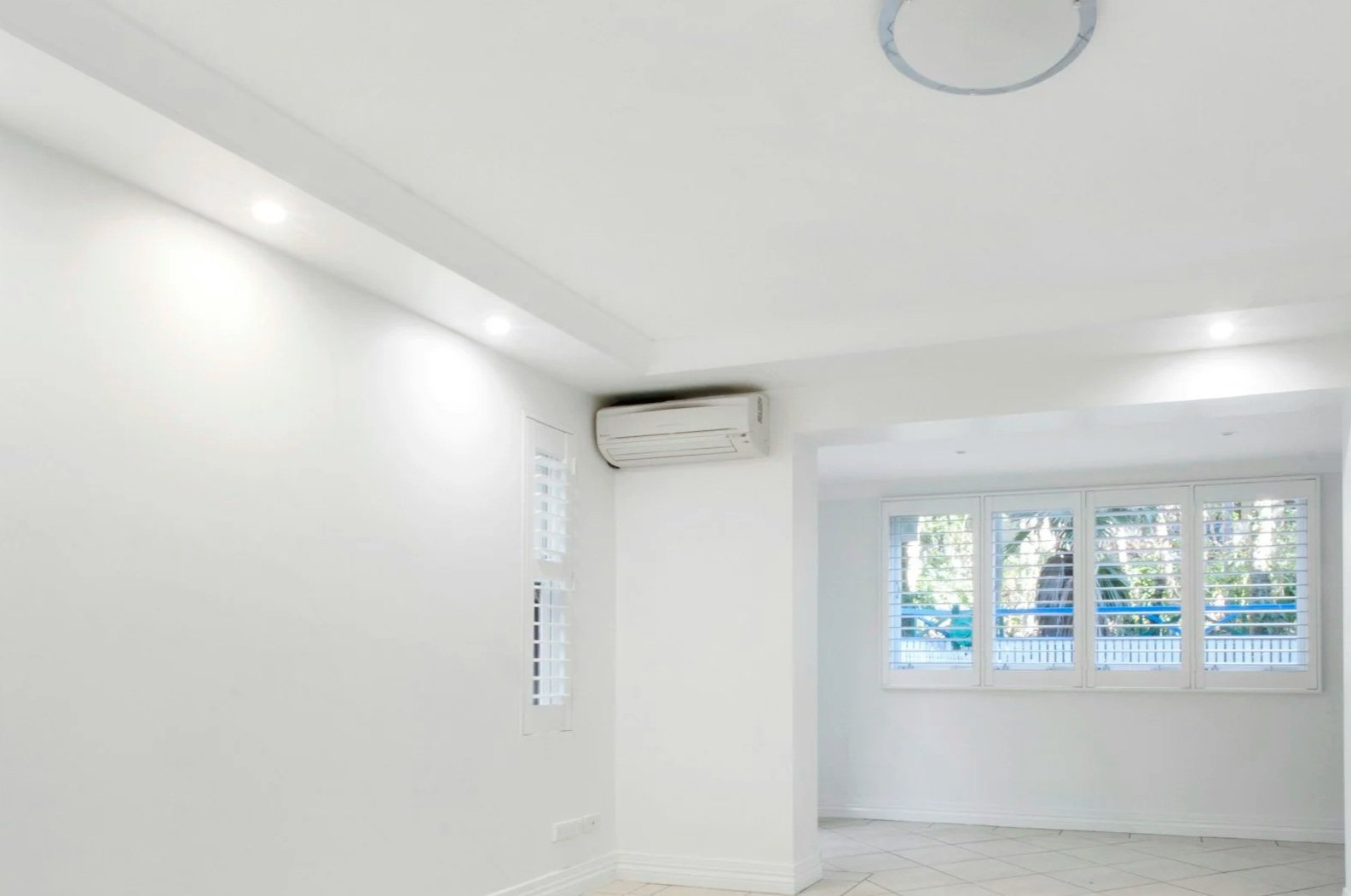 Empty room with white walls, ceiling with recessed lighting, a window with plantation shutters, and an air conditioning unit.