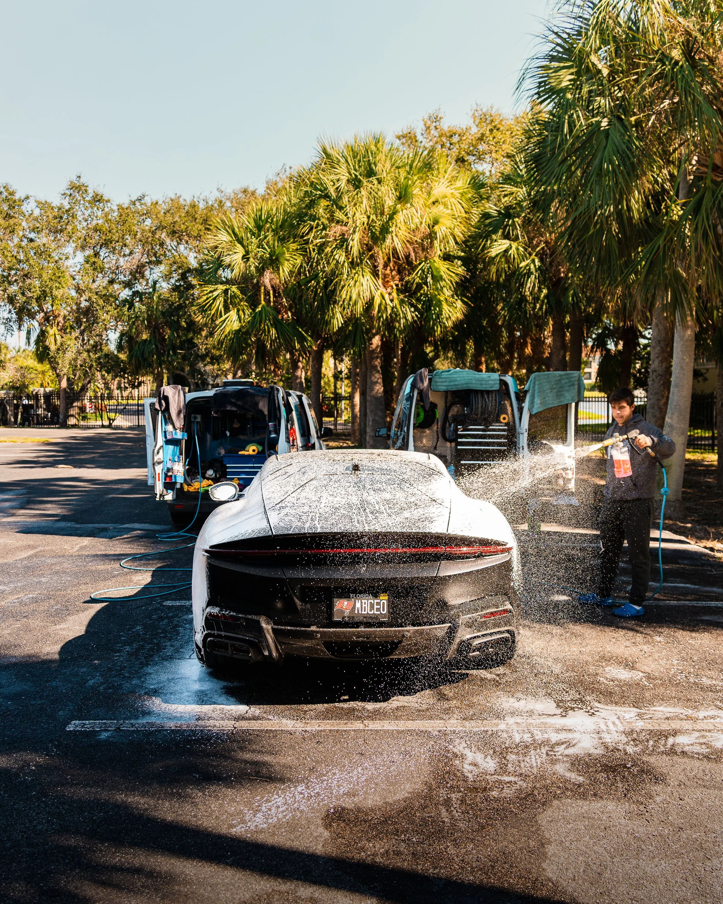 A person washing a sports car with a hose at a car wash station, with two open service vans and palm trees in the background.