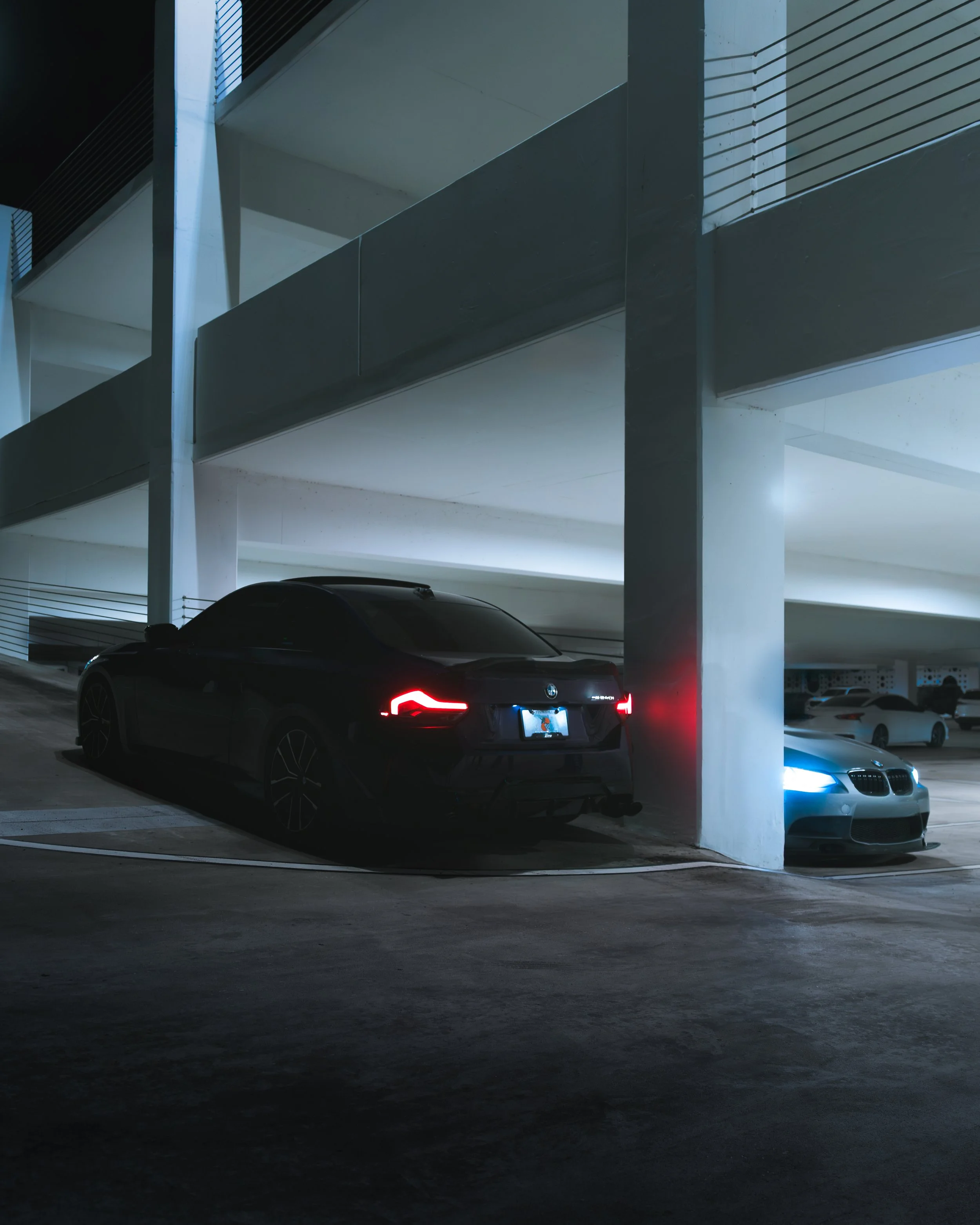 Two cars parked in a multi-level parking garage at night, one black BMW and one silver BMW with blue headlights, with concrete pillars and modern architectural design.