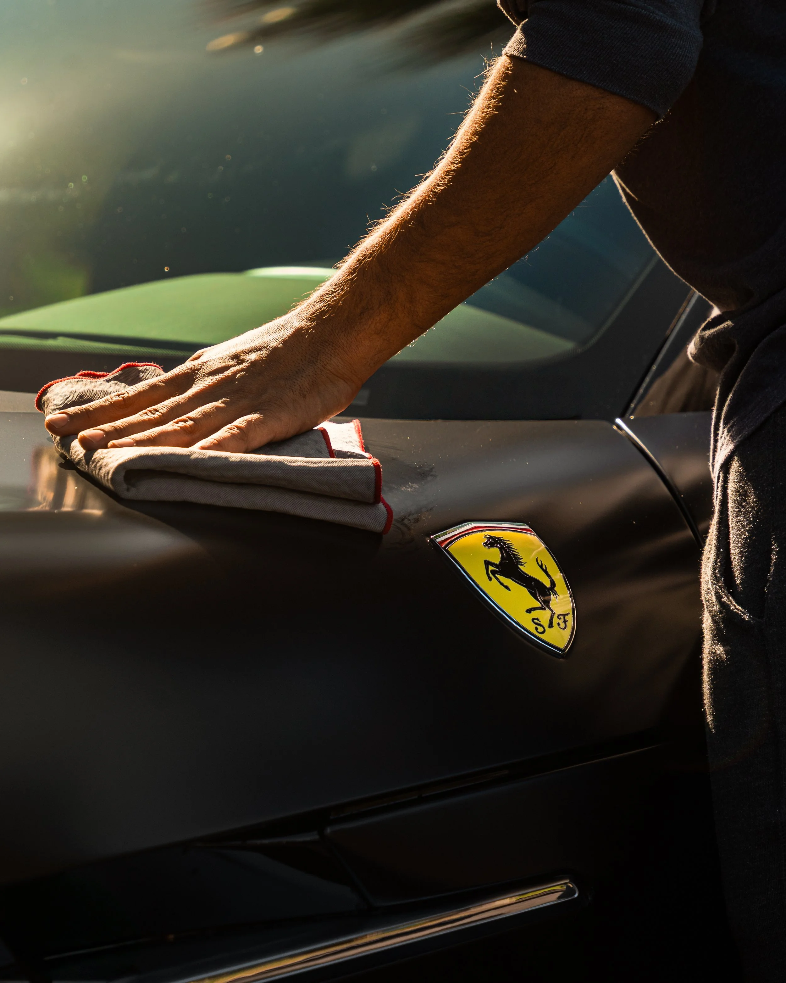 Person polishing the hood of a black Ferrari sports car with a cloth in sunlight