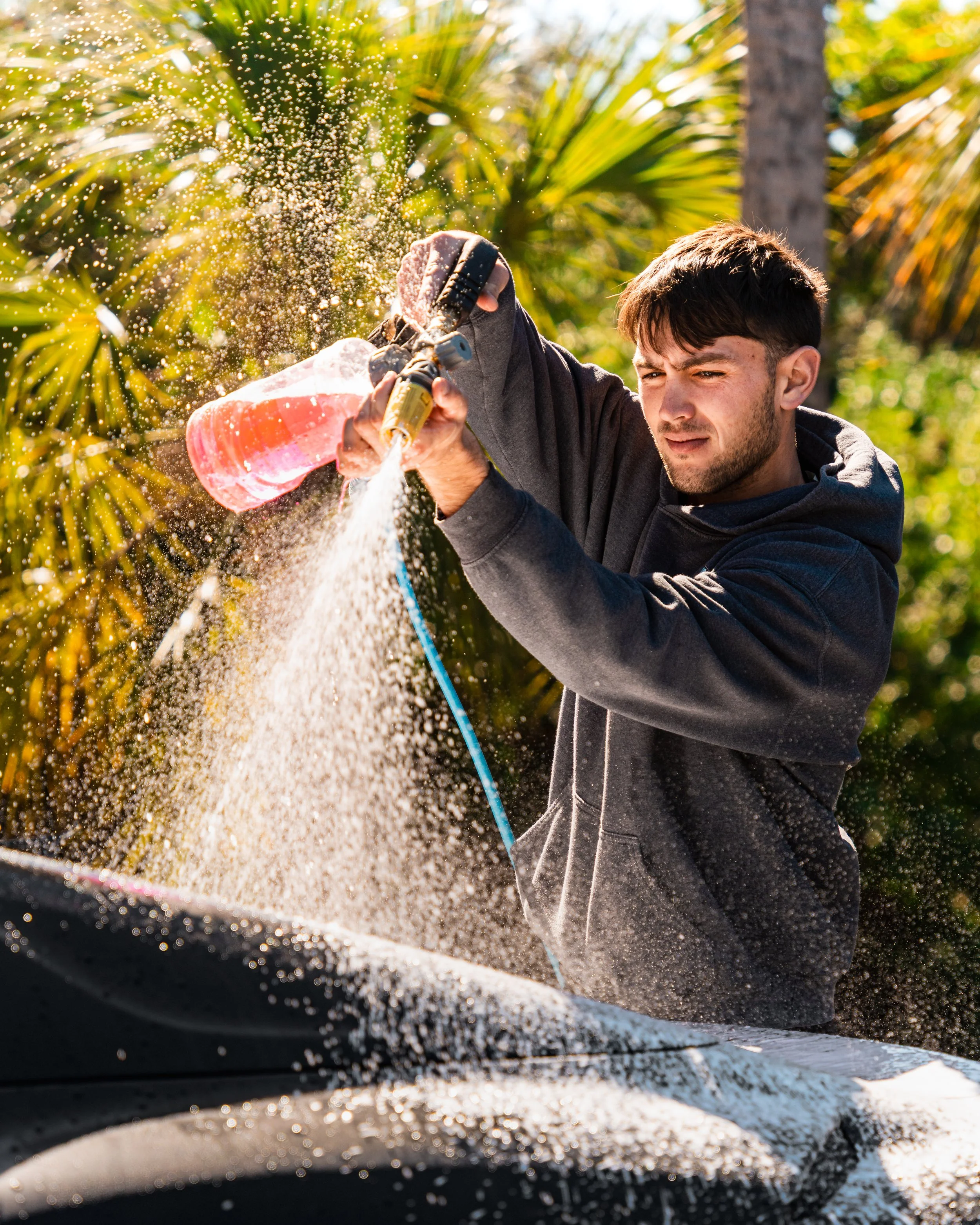 A man wearing a black hoodie washing a black car with a pink soap bottle outdoors amid green palm trees.