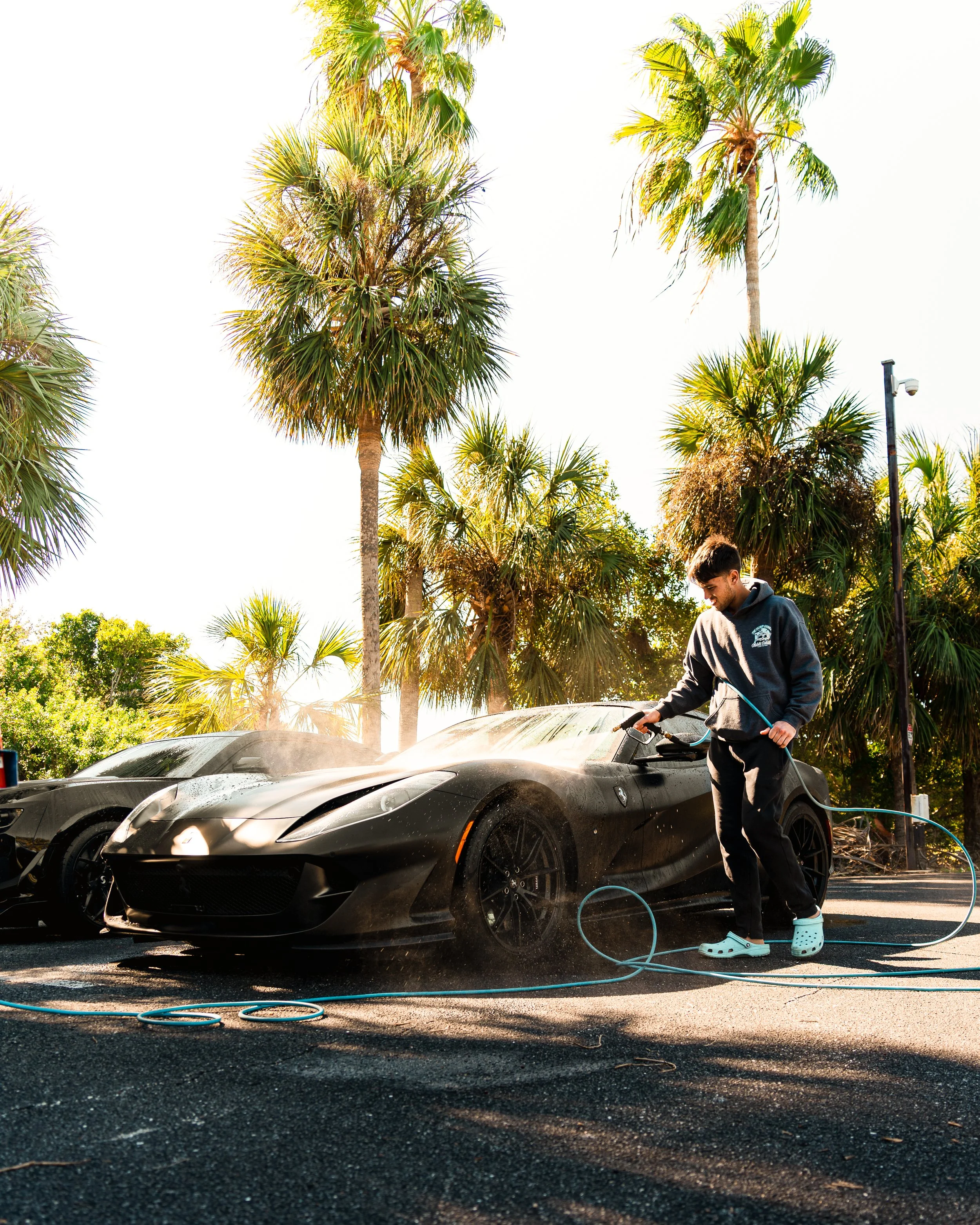 A person washing a black sports car with a hose in an outdoor parking lot surrounded by palm trees on a sunny day.