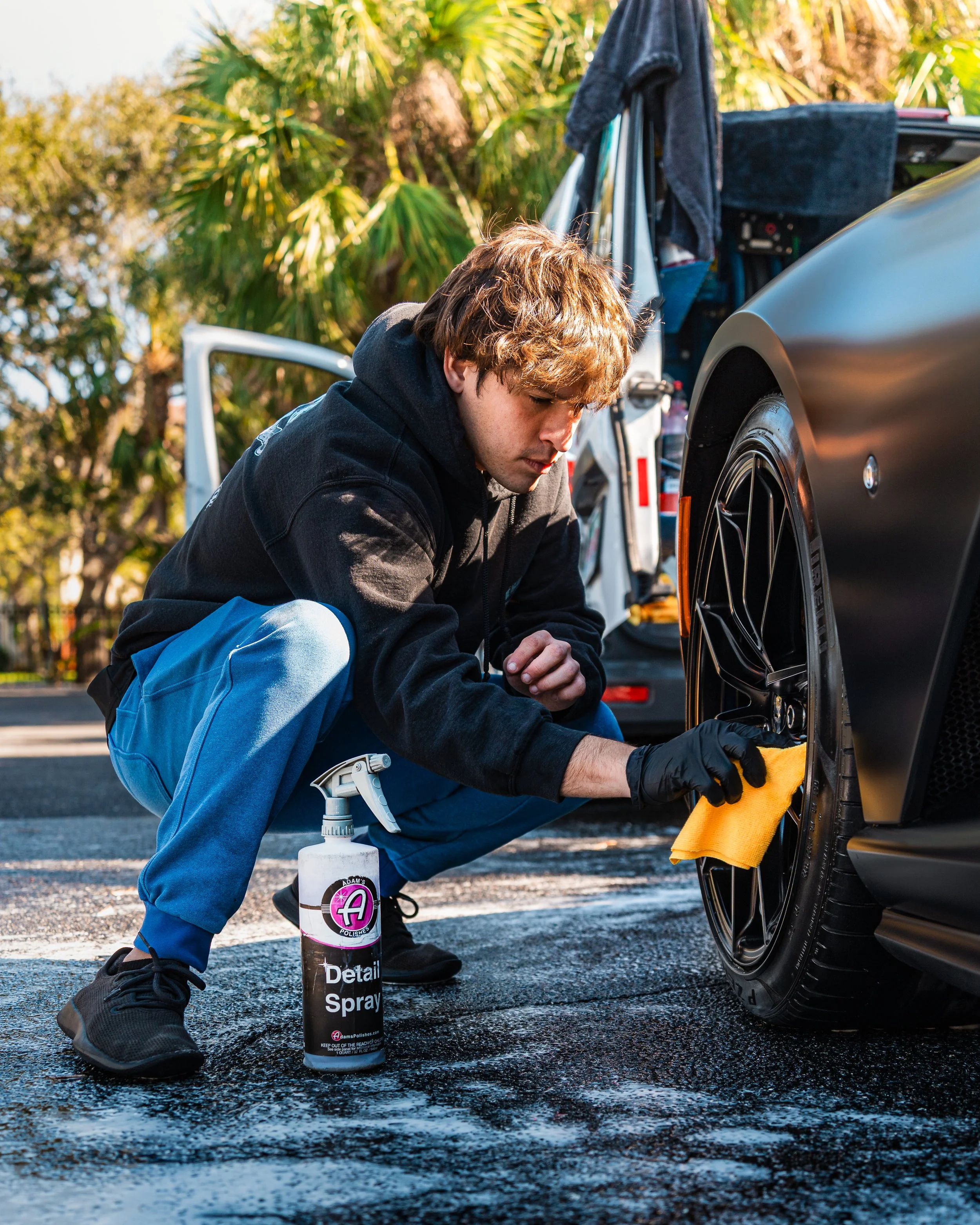 Young man wearing black hoodie and blue pants cleaning the rim of a black sports car with a yellow cloth, surrounded by trees and parked vehicles.