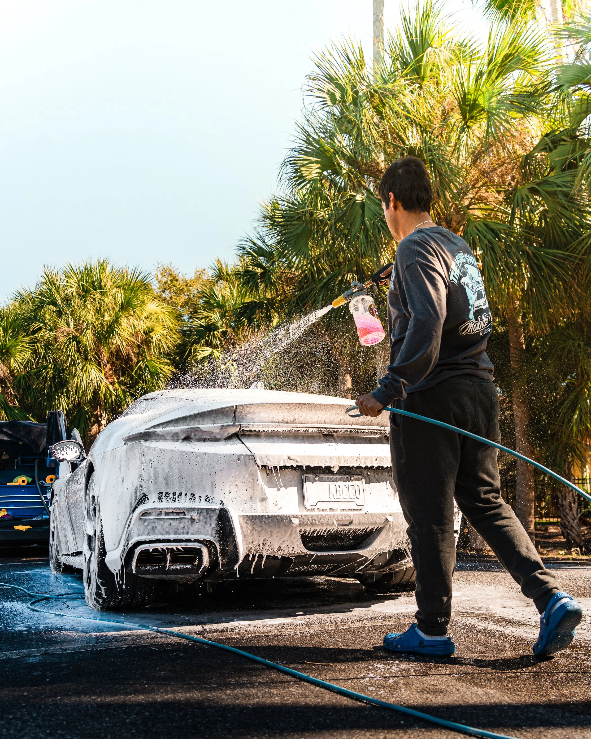 Man washing a white sports car with soap in an outdoor car wash station surrounded by palm trees.