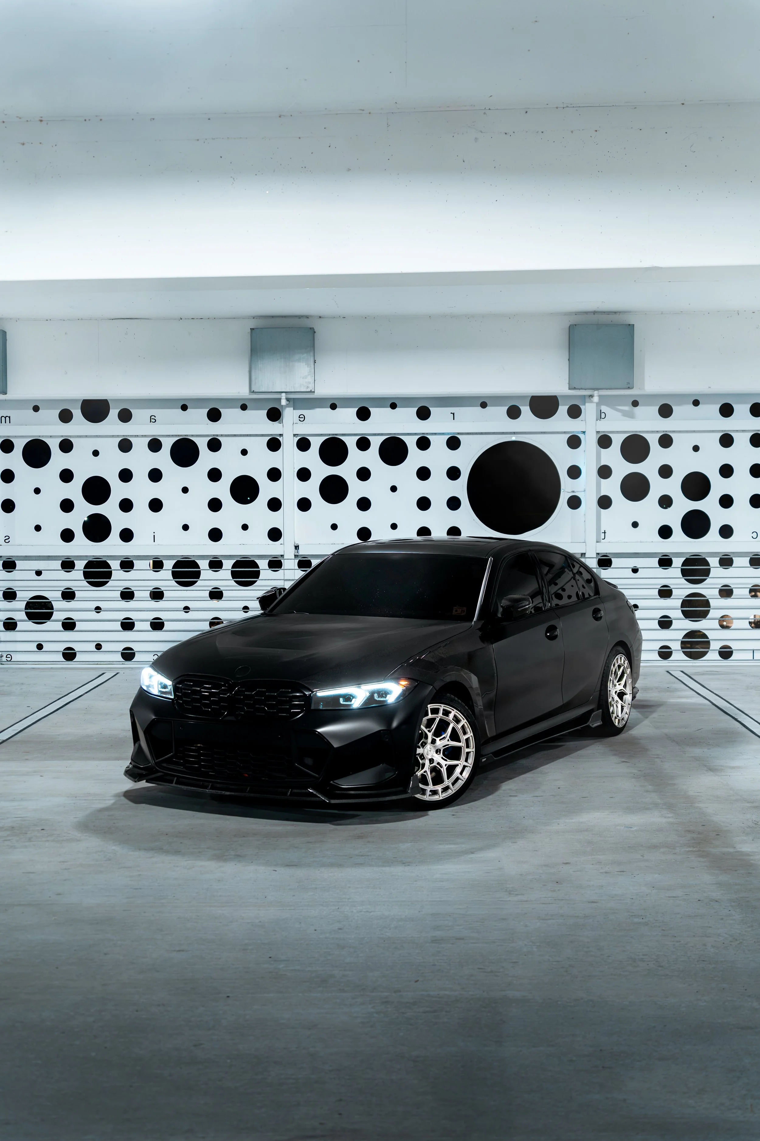 A black sedan car with LED headlights parked in an underground parking garage with a polka dot design on the wall behind it.