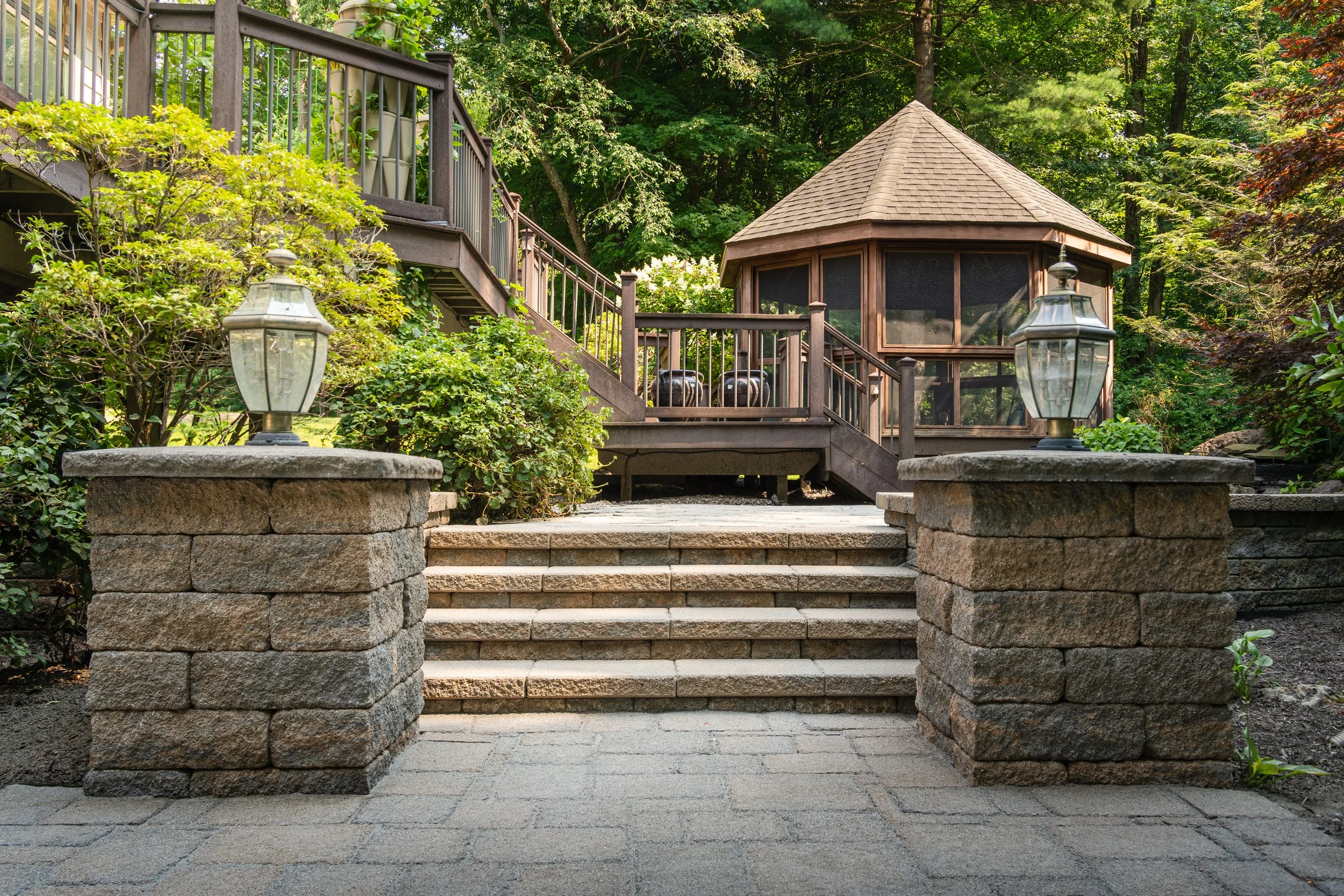 Stone steps leading up to a wooden deck with a gazebo, surrounded by lush green trees and foliage, outdoor lighting fixtures on stone pedestals.