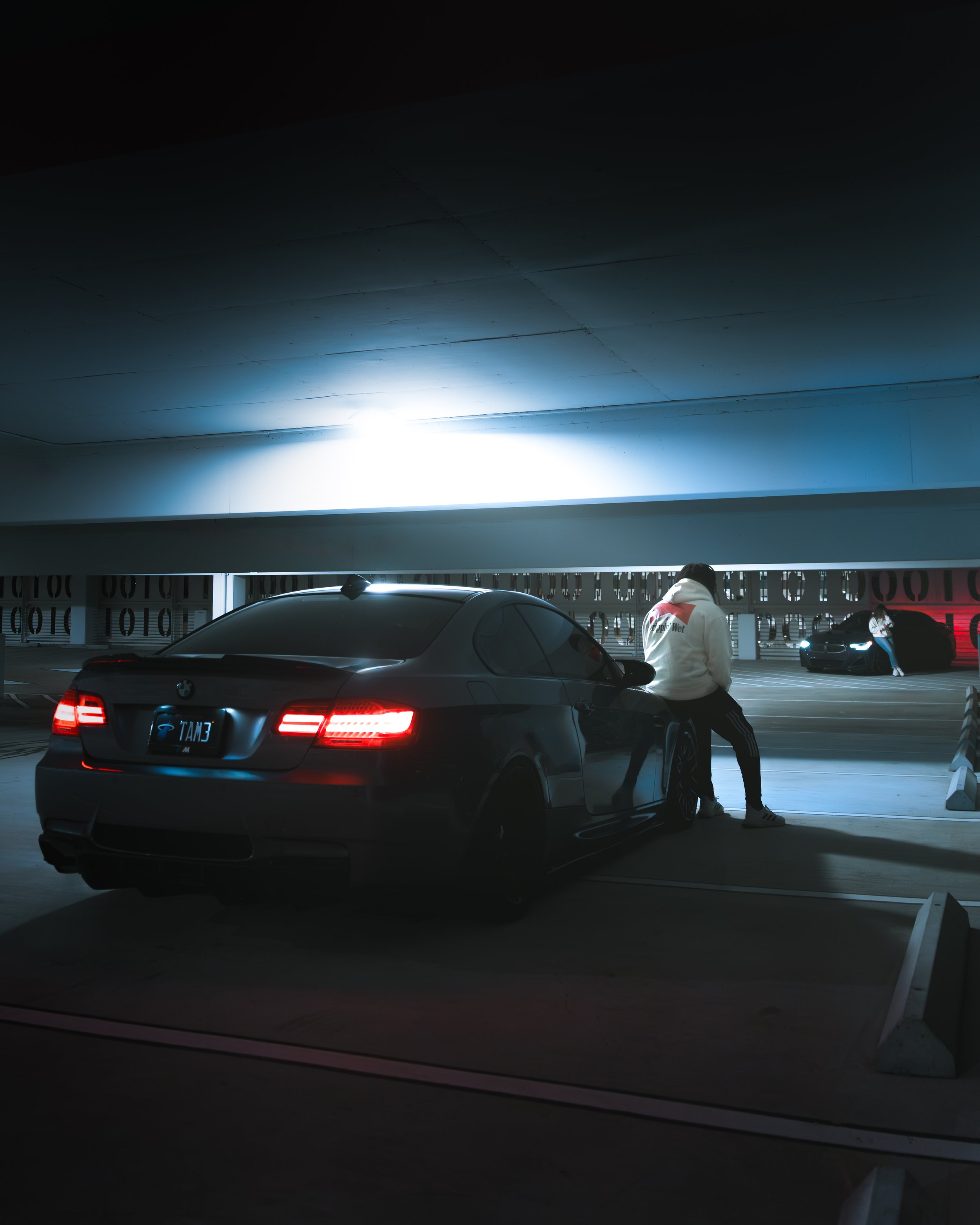 A black BMW parked in a dimly lit underground parking garage with a person standing beside it, illuminated by overhead lights.