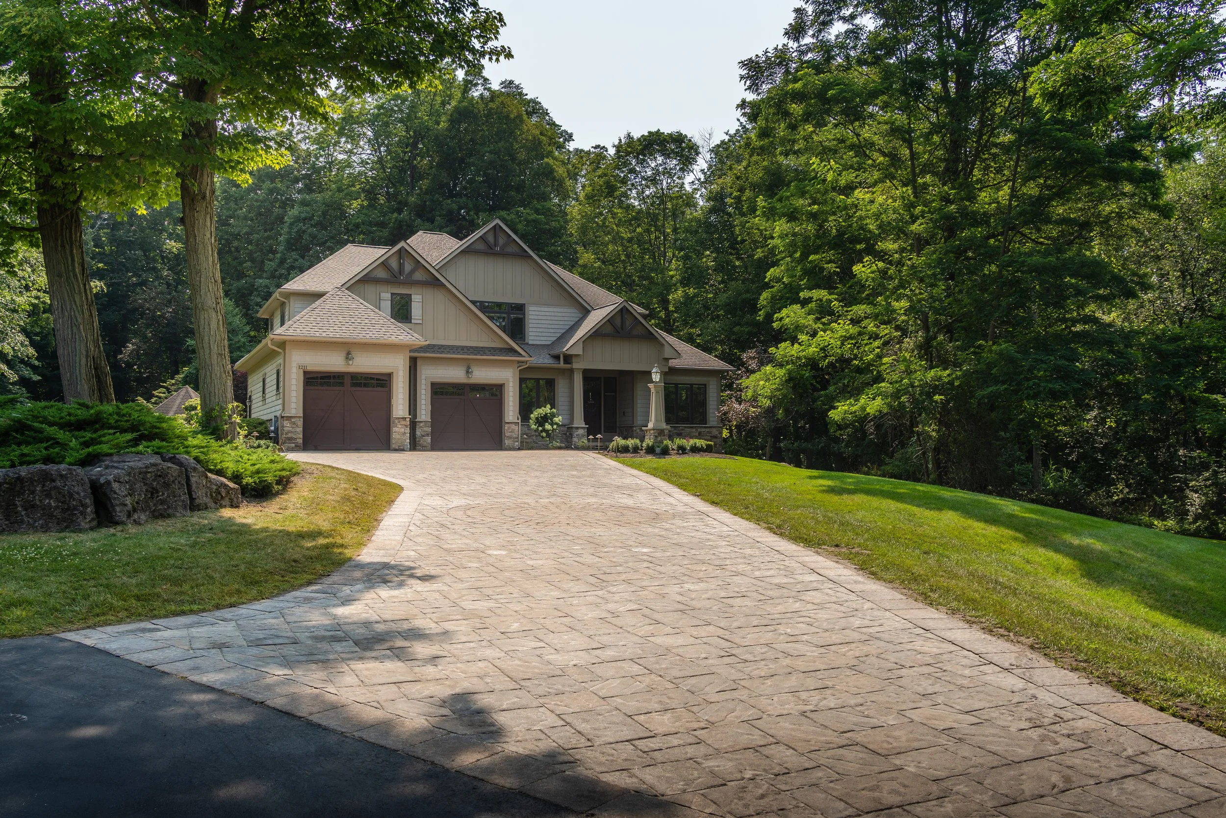 A house with a stone and wood exterior, three garage doors, surrounded by lush green trees and a well-maintained front yard, with a paved driveway leading up to the house.