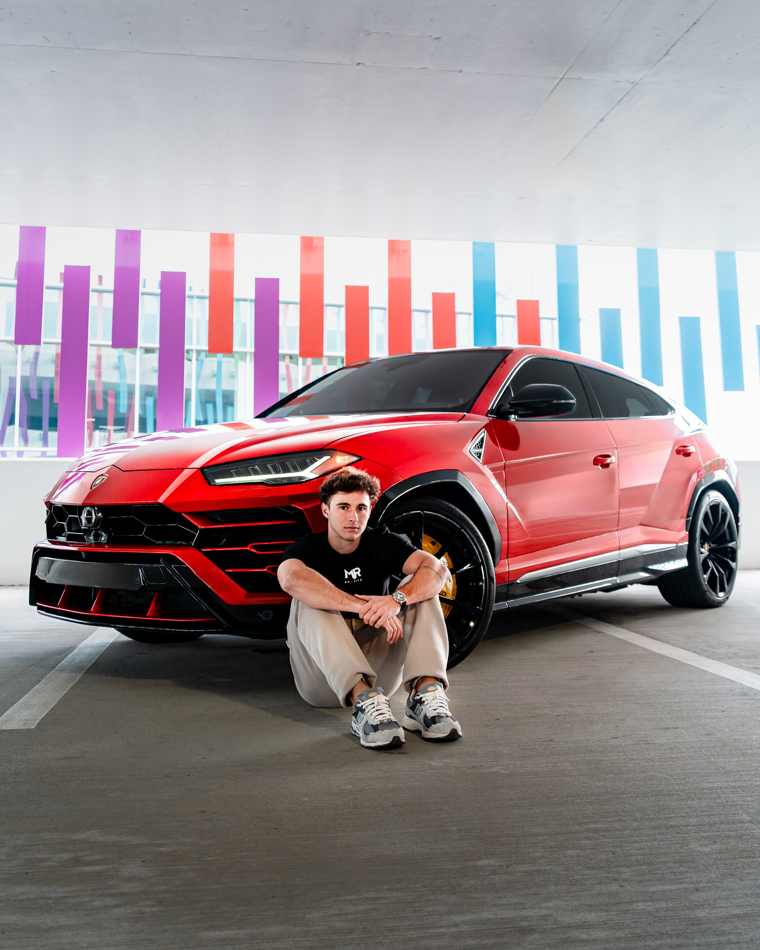 A man sitting on the ground in front of a red sports car in a parking garage with colorful vertical light strips on the wall behind.