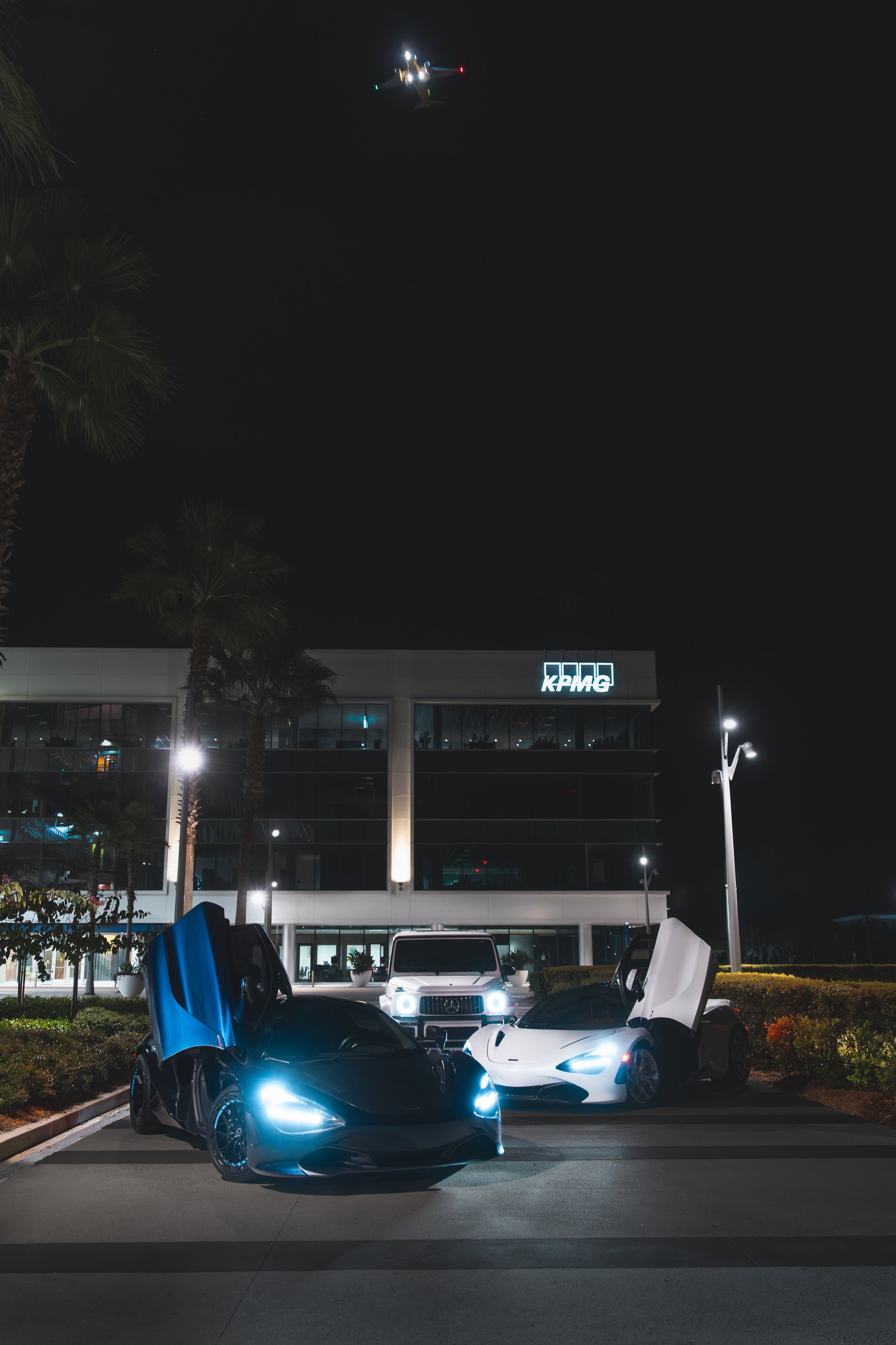 Nighttime scene with two luxury cars with open gullwing doors, a white SUV behind them, and a corporate building with a KPMG logo in the background, illuminated by streetlights, with palm trees and a helicopter flying overhead.