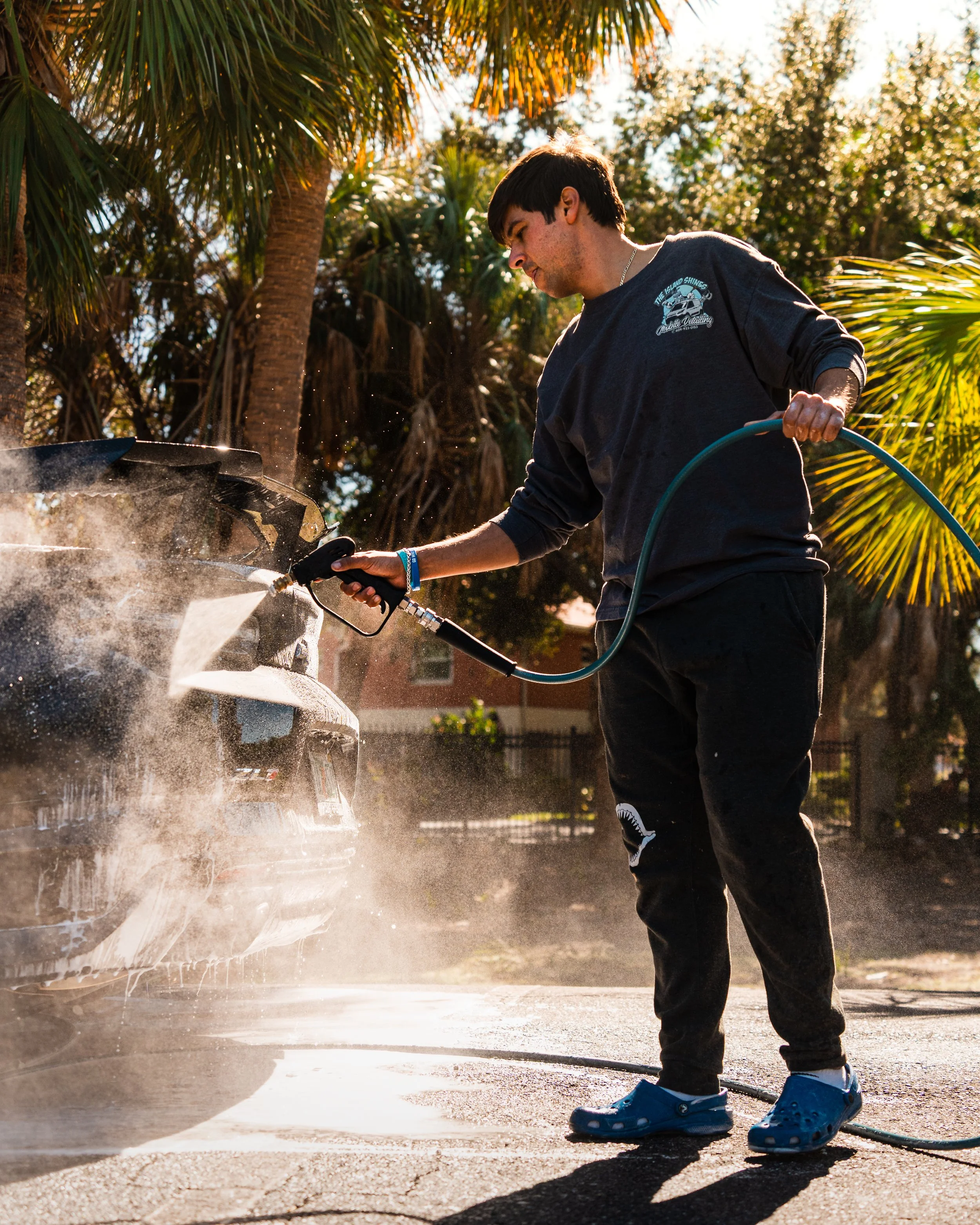 A young man washing a car with a hose in a sunny outdoor setting with palm trees in the background.