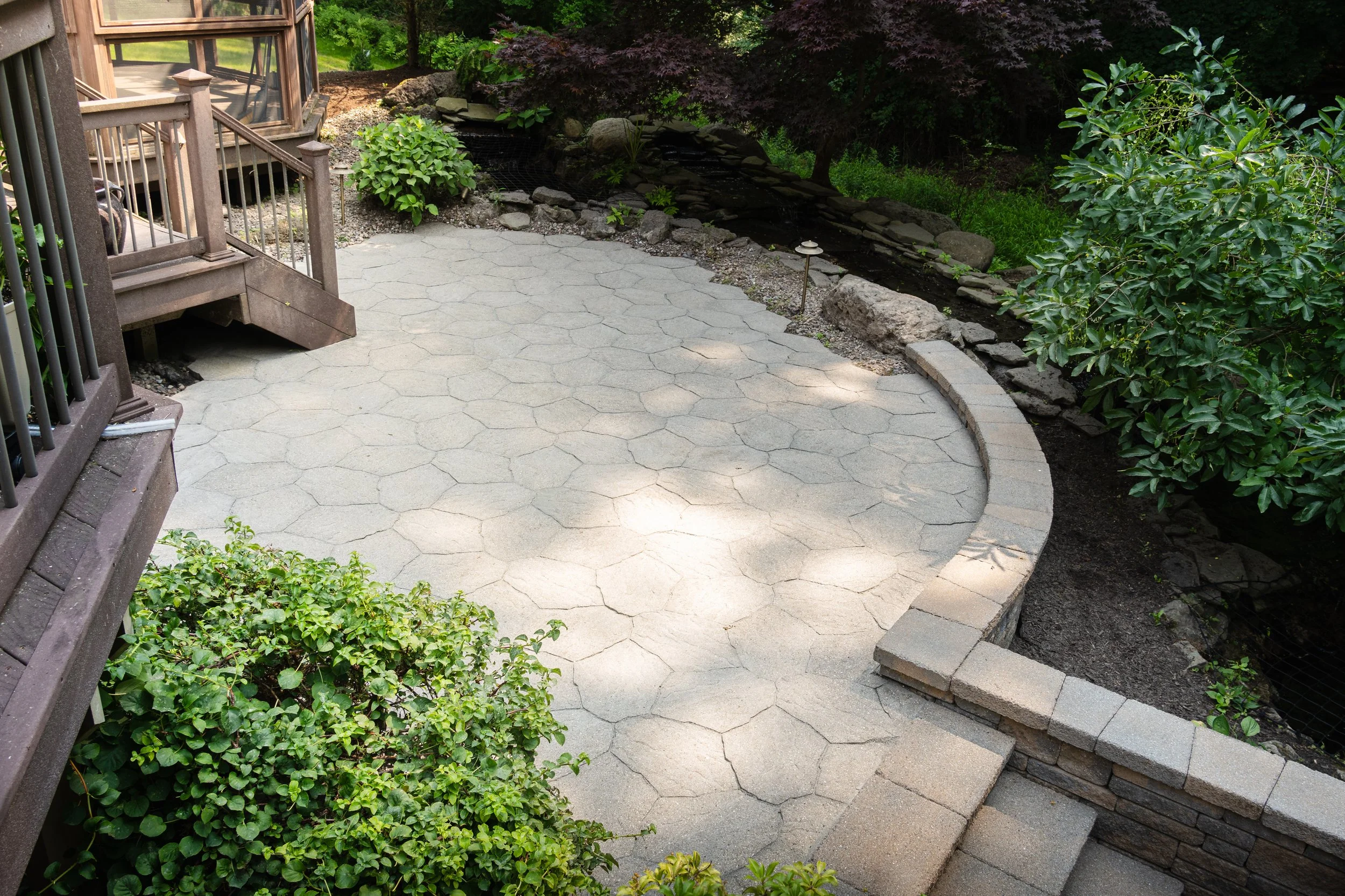 A backyard patio with a curved stone wall, lush green bushes, a small black water feature, and a deck with stairs leading down.