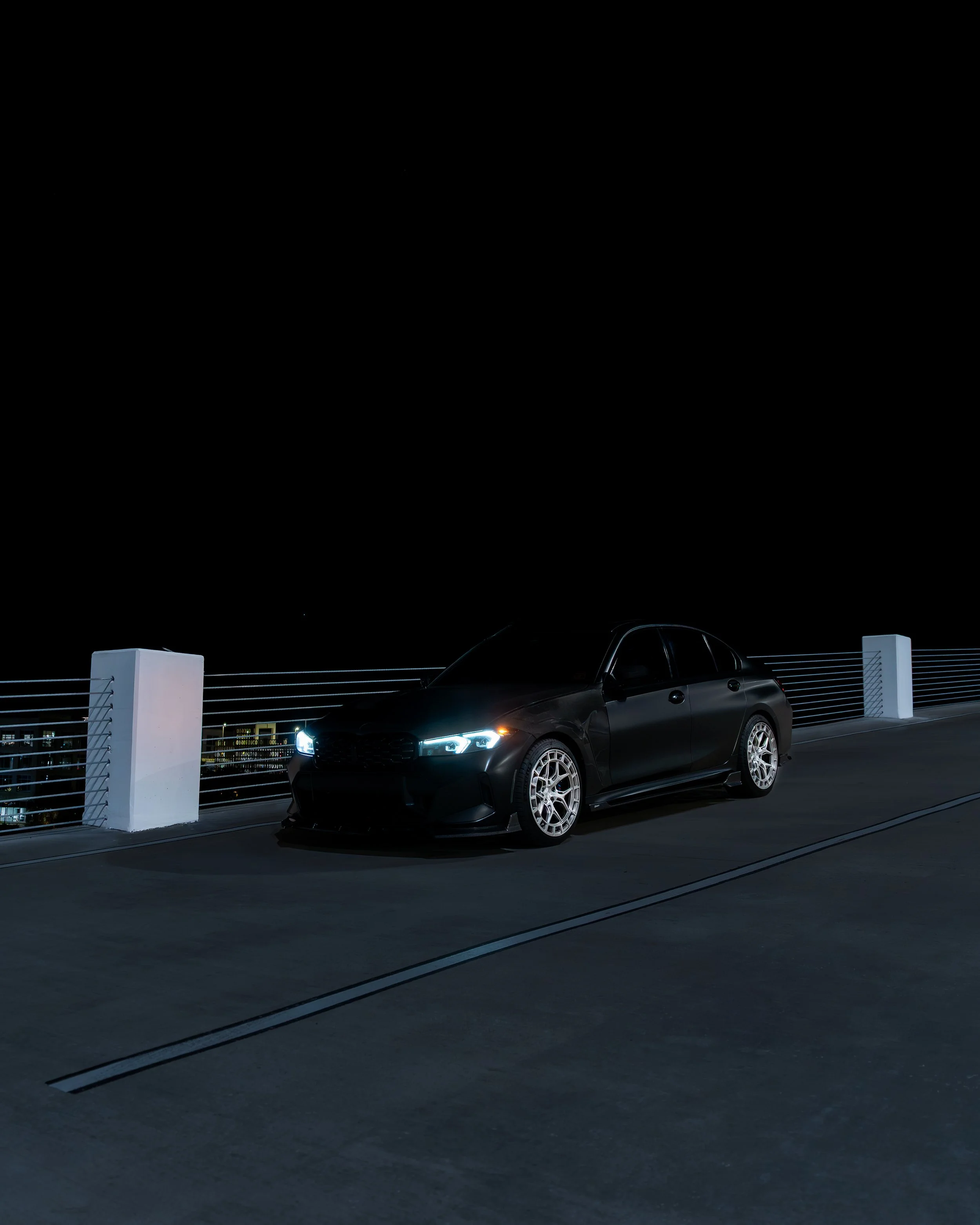 A black sedan with illuminated headlights parked on a rooftop at night, with city lights in the background.