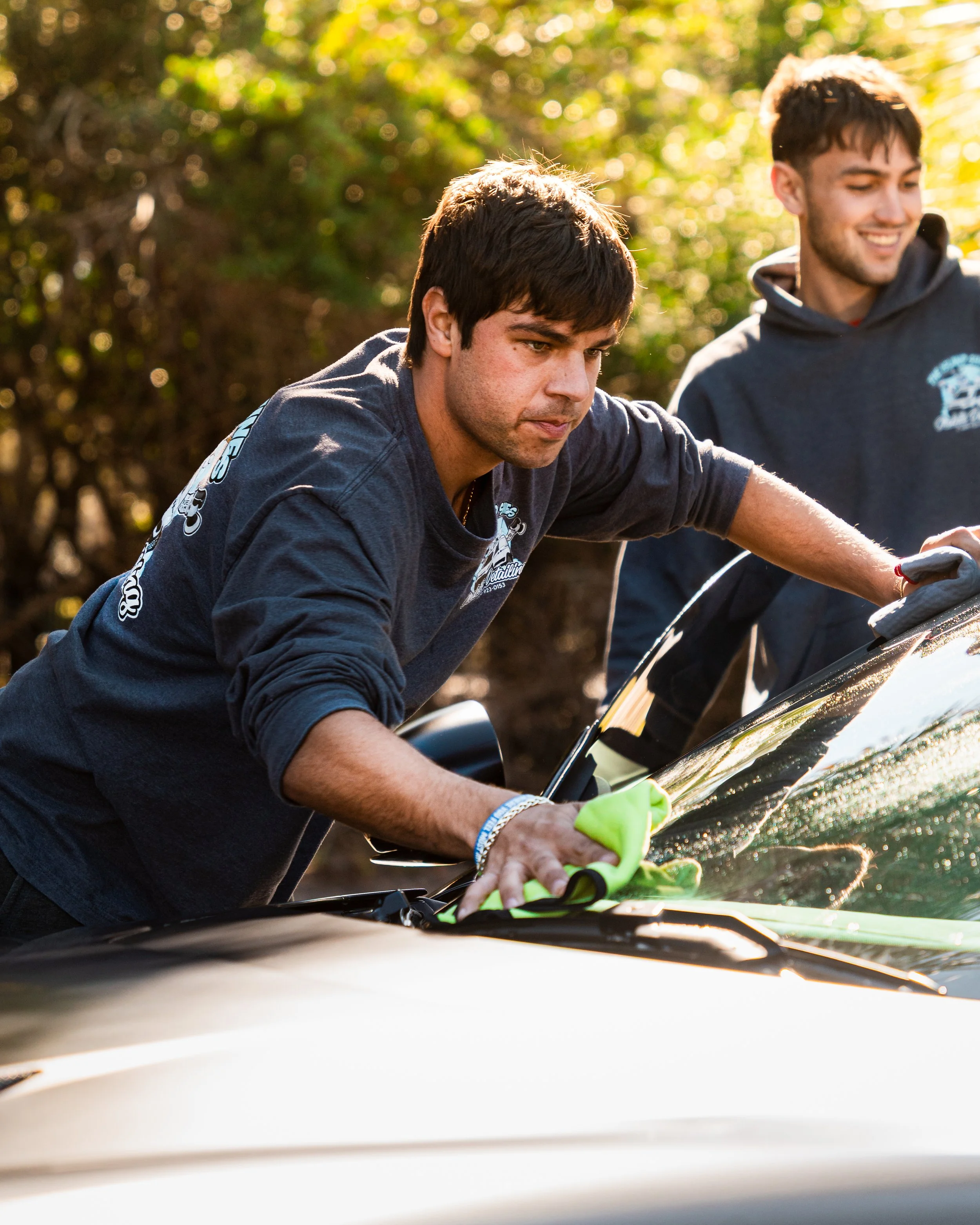 Two young men cleaning or repairing a black car outdoors during daytime, one is wiping the windshield with a cloth, both wearing dark sweatshirts.